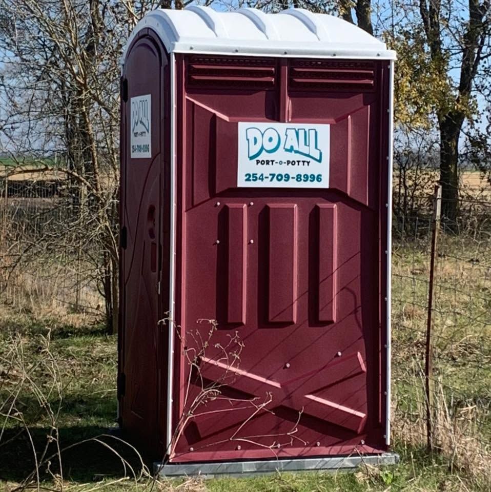 Maroon portable toilet with white roof and Do All sign, standing in a grassy field.