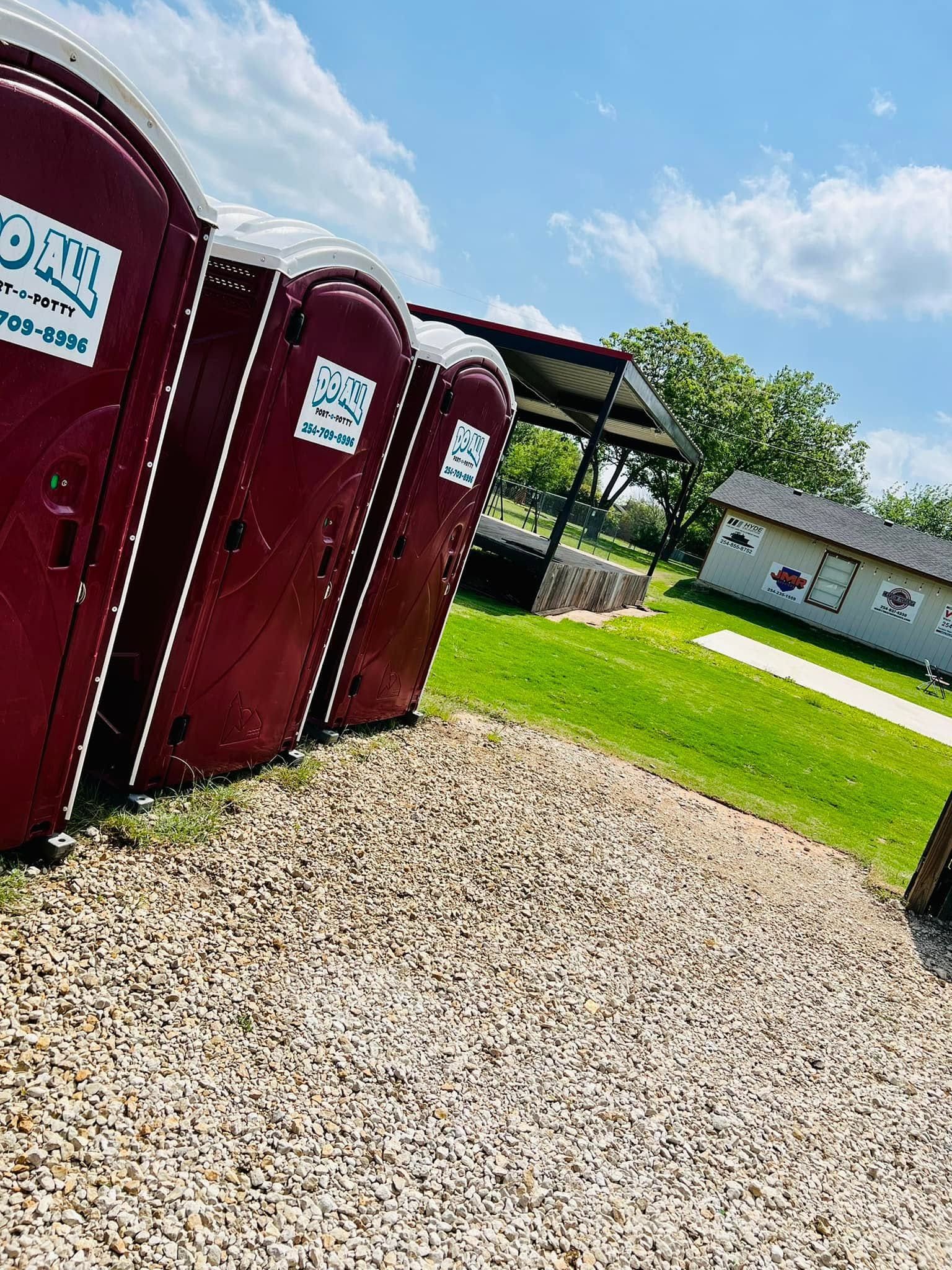 Row of maroon portable toilets beside a gravel path, with a grassy yard, pavilion, and house in the background