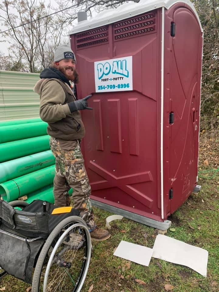 Worker beside red portable toilet on muddy construction site with green pipes and a wheelchair nearby