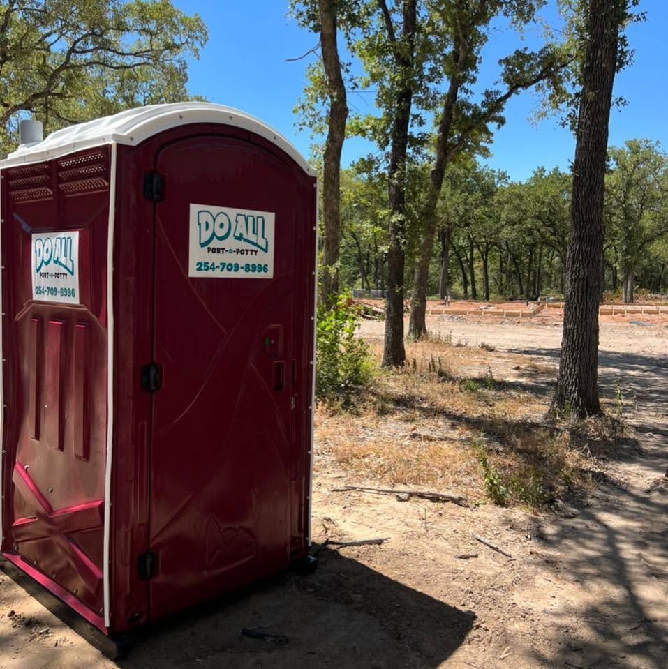 Maroon portable toilet in a wooded outdoor area with a “Do All” sign on the door