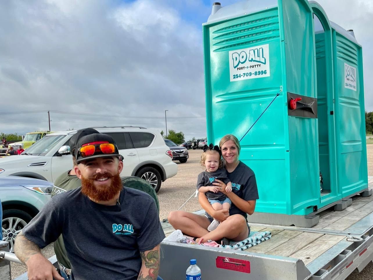 Two people sitting on a trailer beside a teal portable toilet in a parking lot