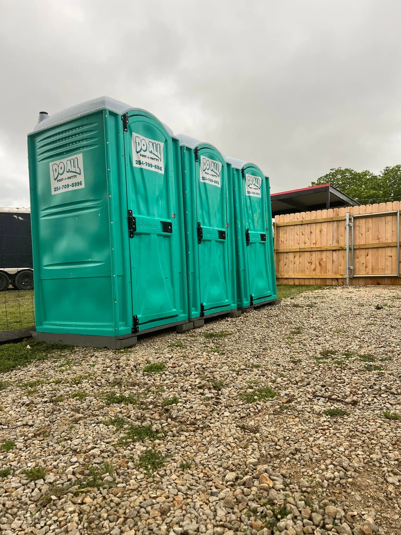 Three turquoise portable toilets on gravel beside a wooden fence under a cloudy sky