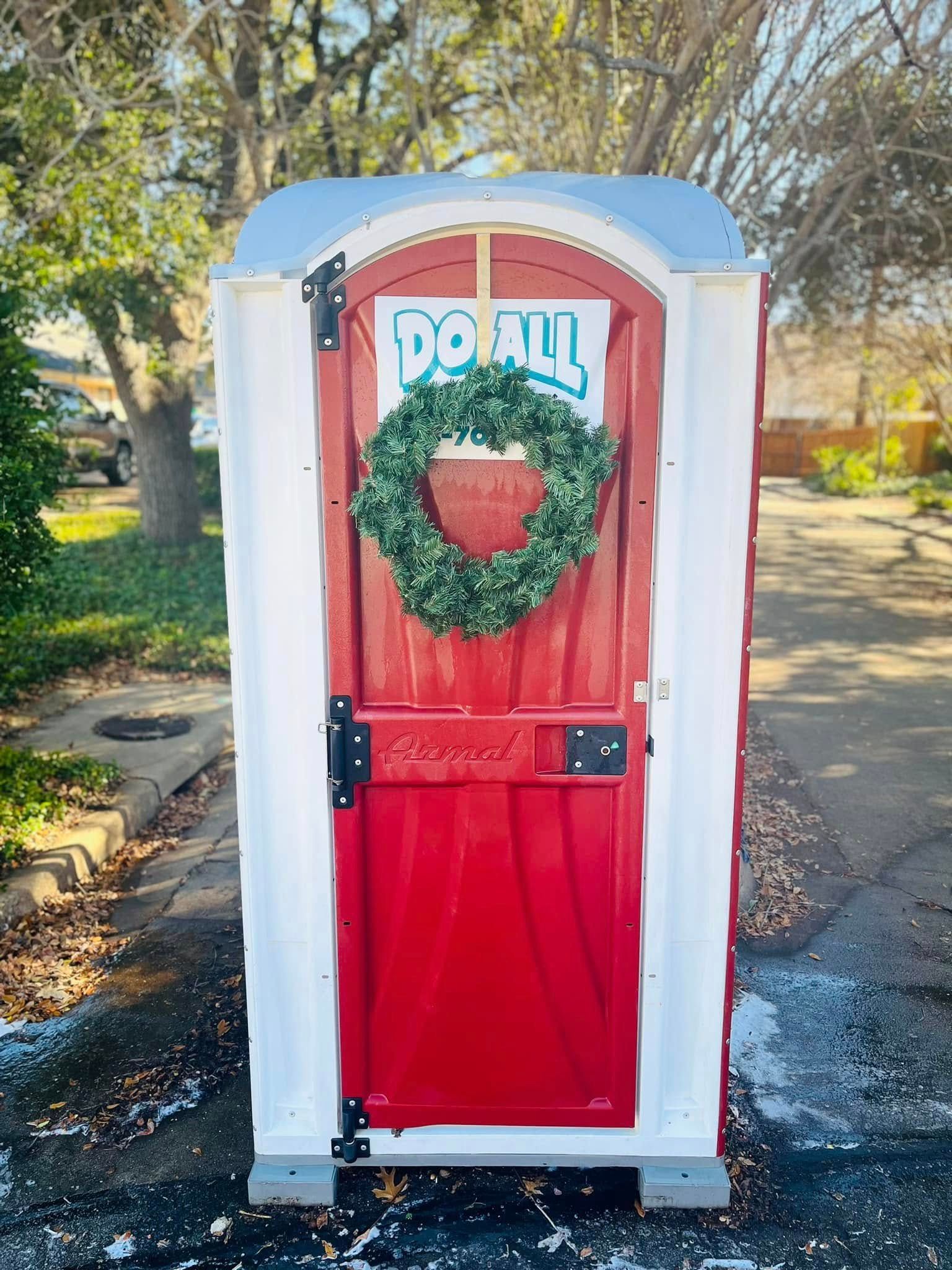 Red portable toilet decorated with a holiday wreath and “Jolly” sign outdoors on a sunny day
