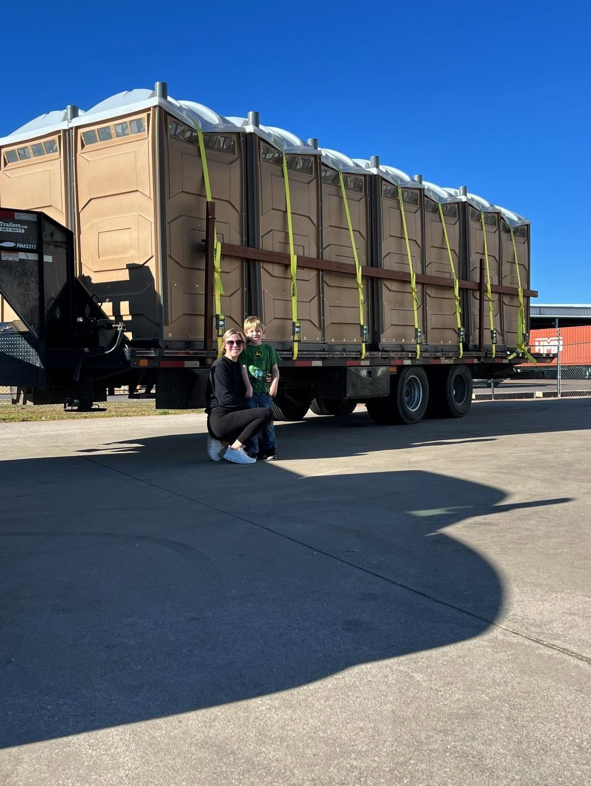 Person crouching beside a large brown tarp-covered trailer in a sunny parking lot.