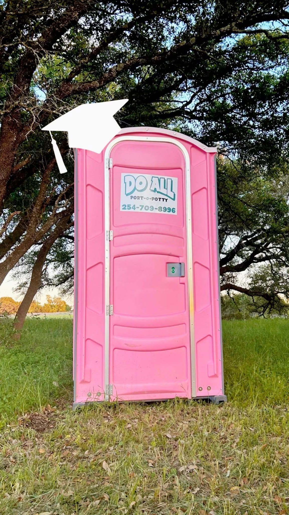 Pink portable toilet in a grassy field with trees in the background.