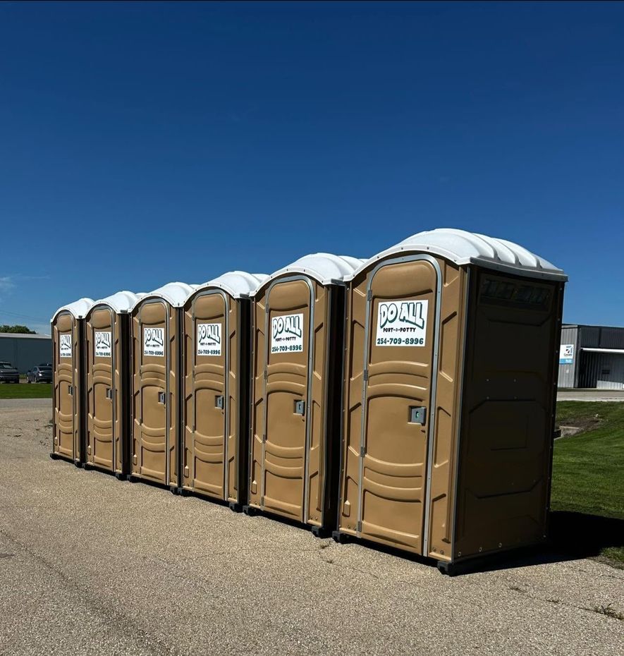 Row of tan portable toilets with snow on top beside a paved road under a clear blue sky