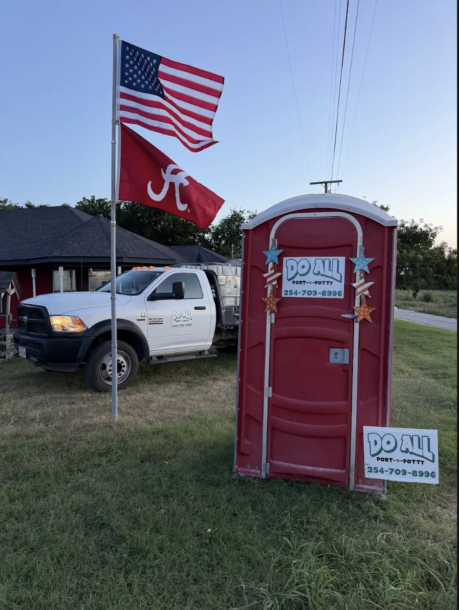 Red portable toilet with Alabama sign, near a white pickup truck and two flags by a roadside.