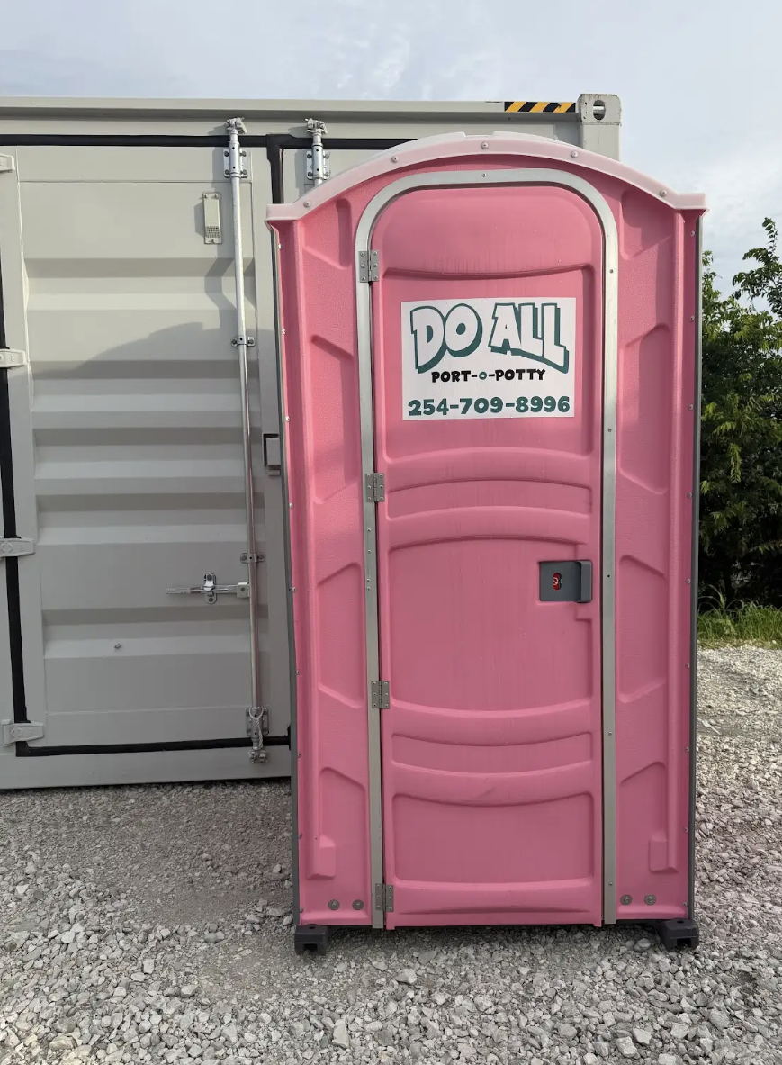 Pink portable toilet with a “Do All” logo in front of a gray shipping container.