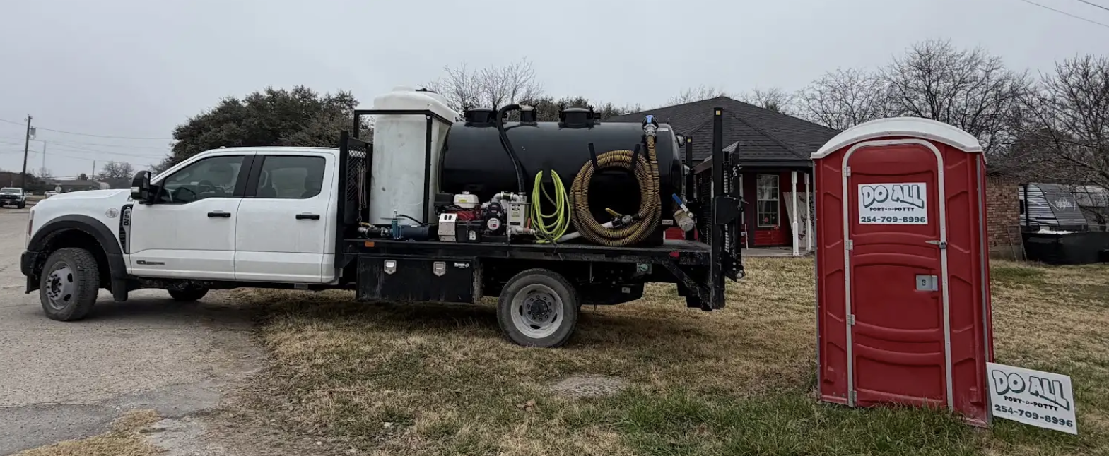 White utility truck with equipment parked beside a red portable toilet on a cloudy day.