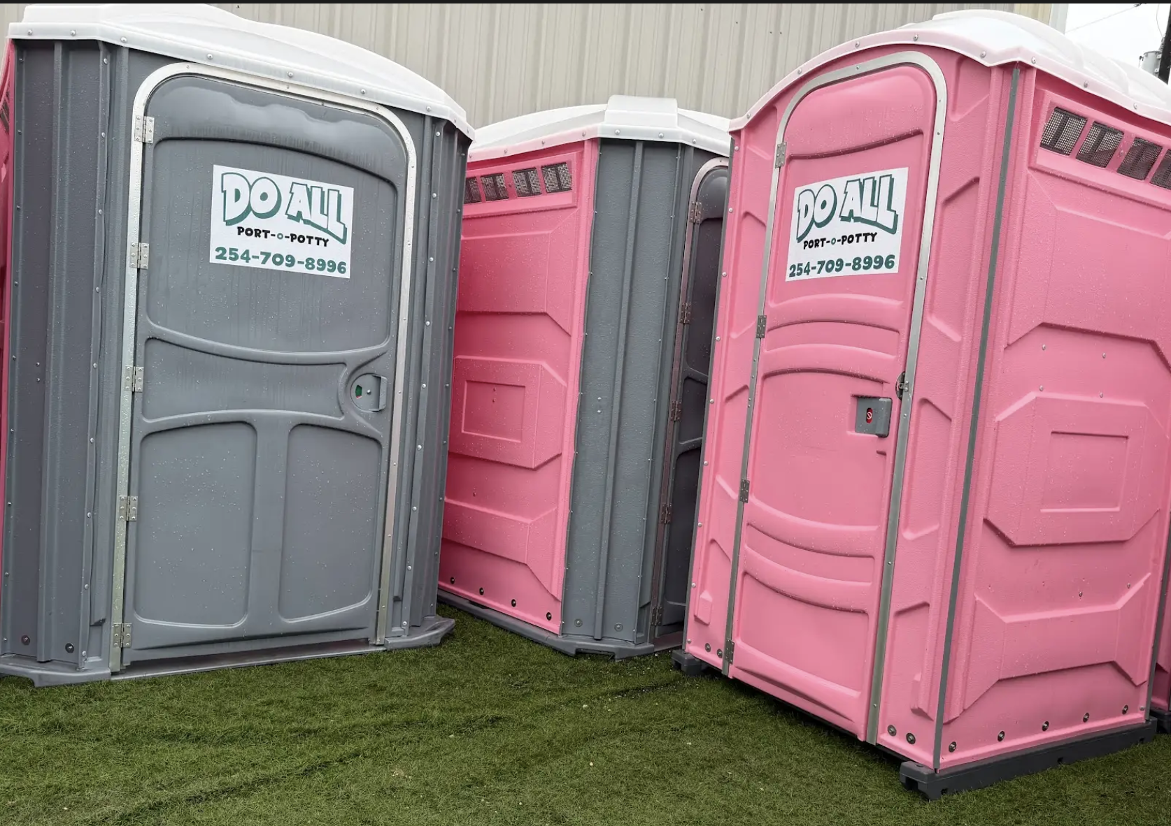 Three portable toilets, two pink and one gray, lined up outdoors on grass.