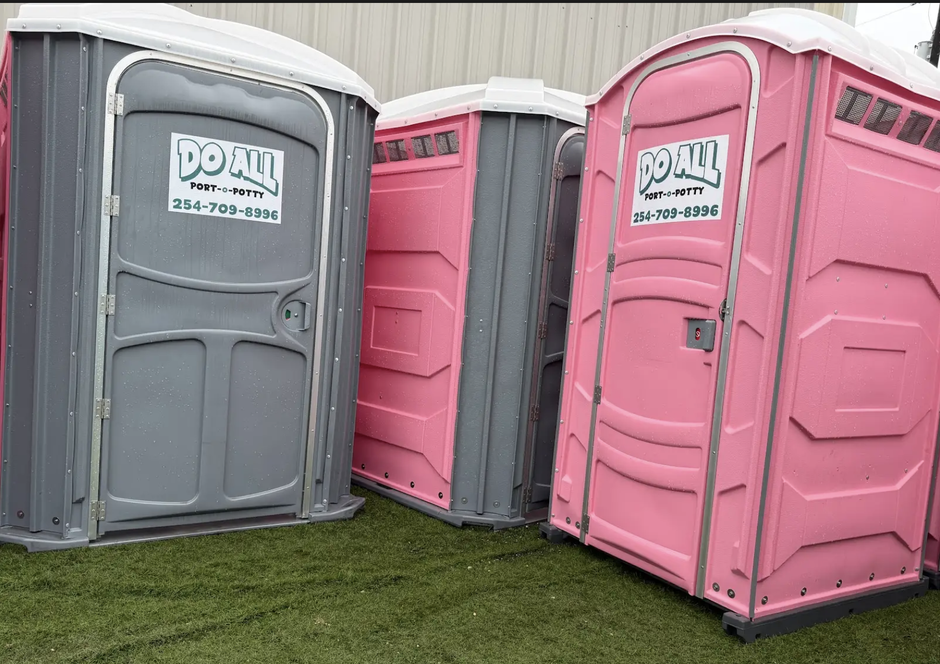 Three portable toilets, two pink and one gray, lined up outdoors on grass.