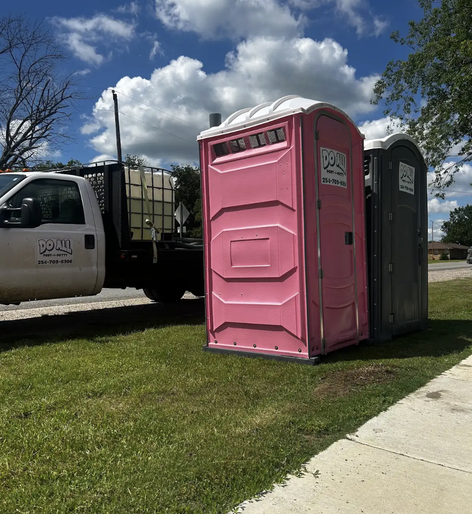 Pink and gray portable toilets beside a truck on a grassy roadside under a blue sky