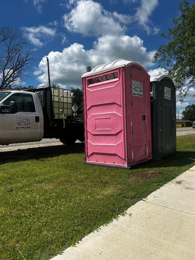 Pink portable toilet beside a dark gray portable toilet on a grassy curb near a truck under a blue sky