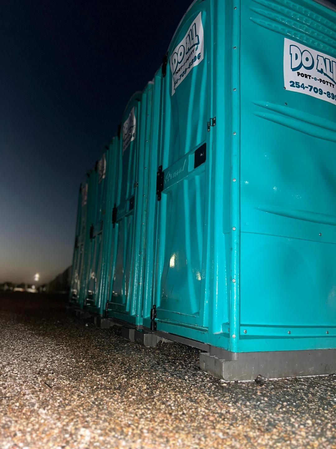 Rows of turquoise portable toilets at night on a gravel lot