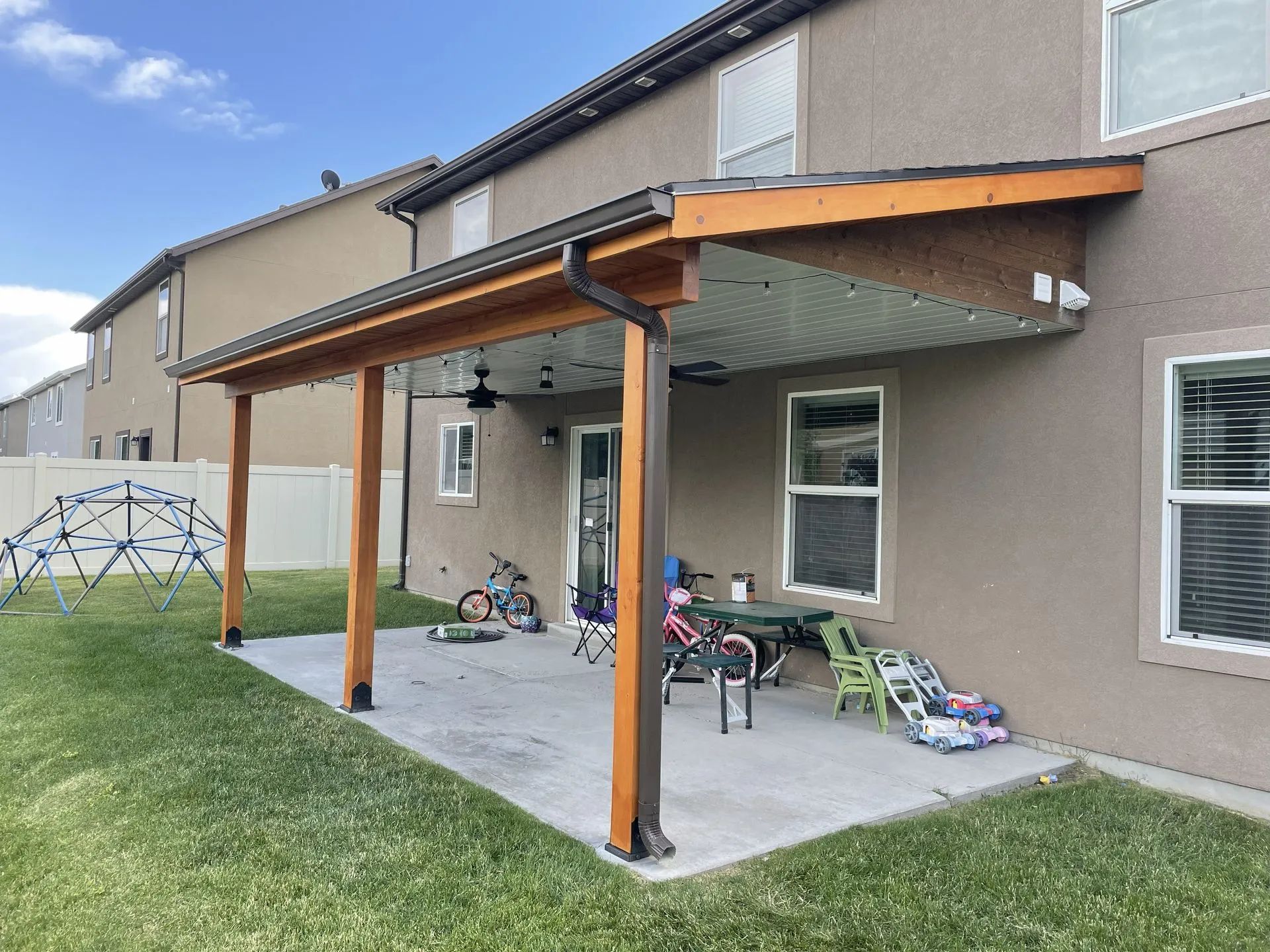 Covered patio with wooden posts beside a beige house, with children’s bikes and toys on the concrete slab.