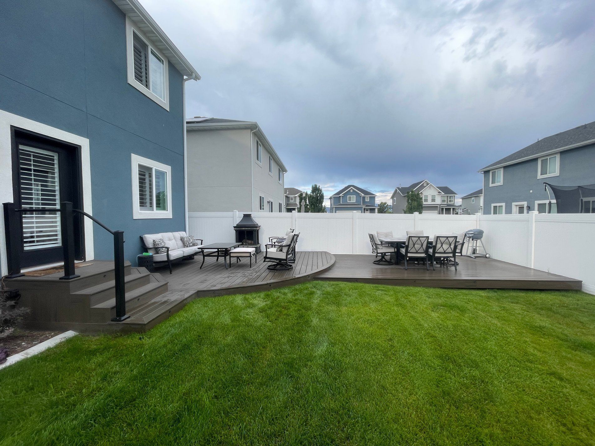 Backyard deck with patio tables and chairs beside a green lawn and blue house under cloudy sky