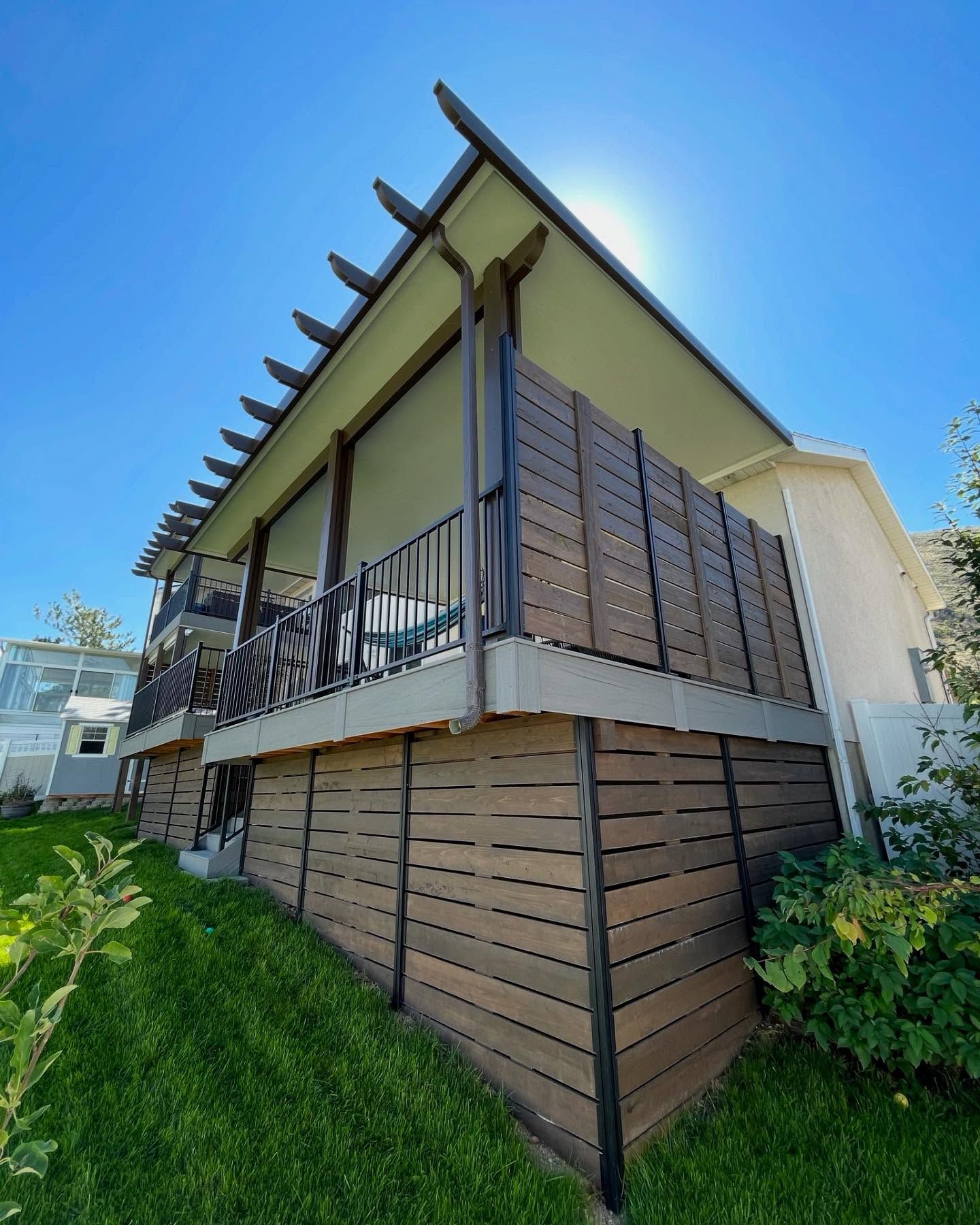 Modern raised house with a covered balcony, wood siding, and green lawn under a bright blue sky