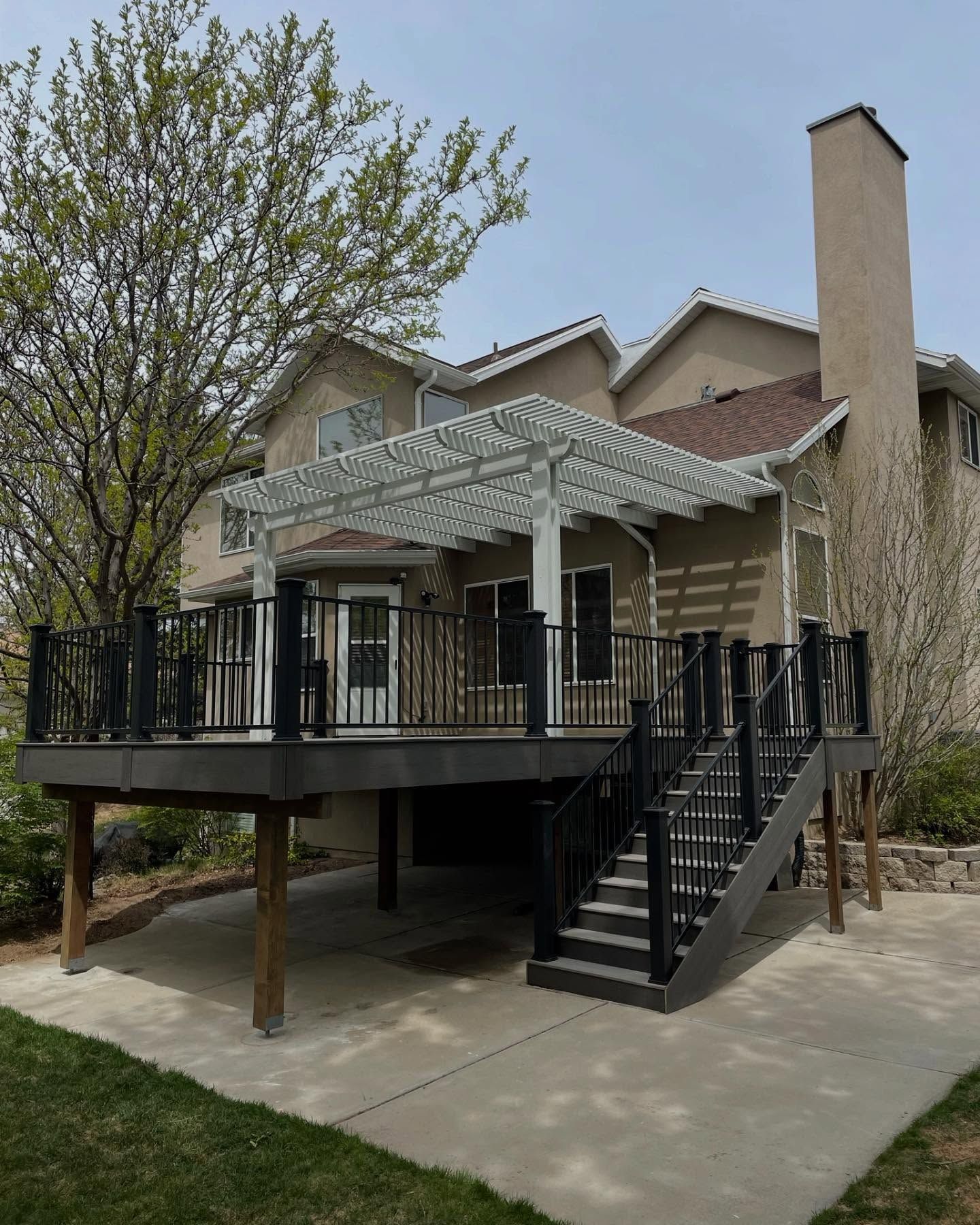 Raised house deck with white pergola, black stairs, and a concrete patio in a suburban yard