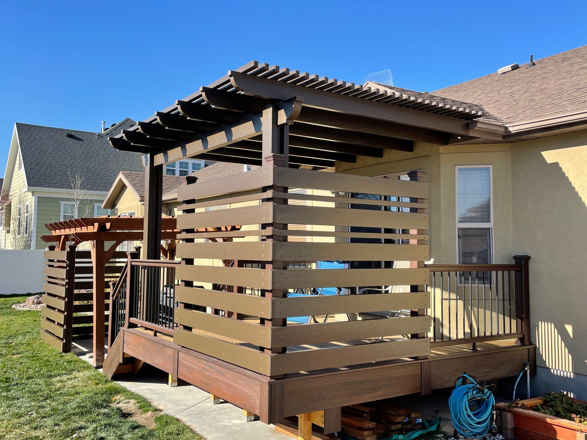 Wooden backyard deck pergola with slatted privacy walls beside a house and patio on a sunny day