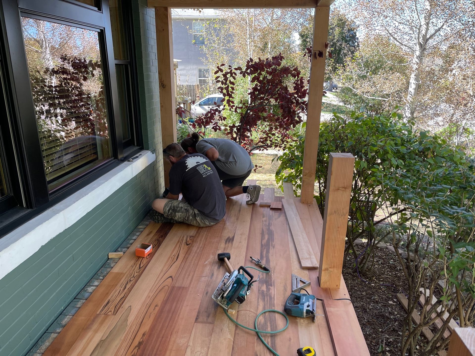 Two workers installing wooden deck boards on a porch, with tools and greenery around them.