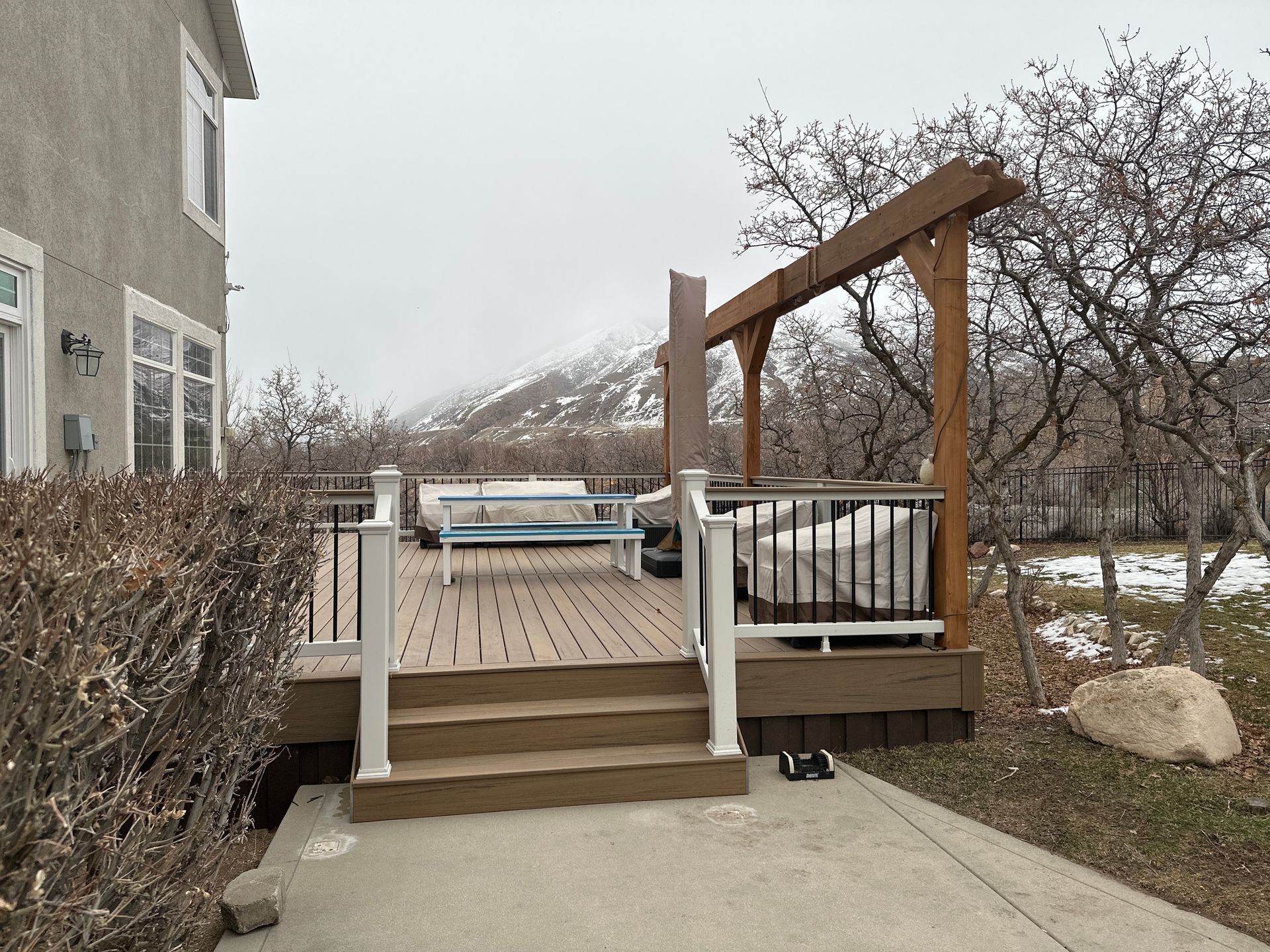 Wooden deck with steps and railing beside a house, overlooking a snowy backyard and trees.