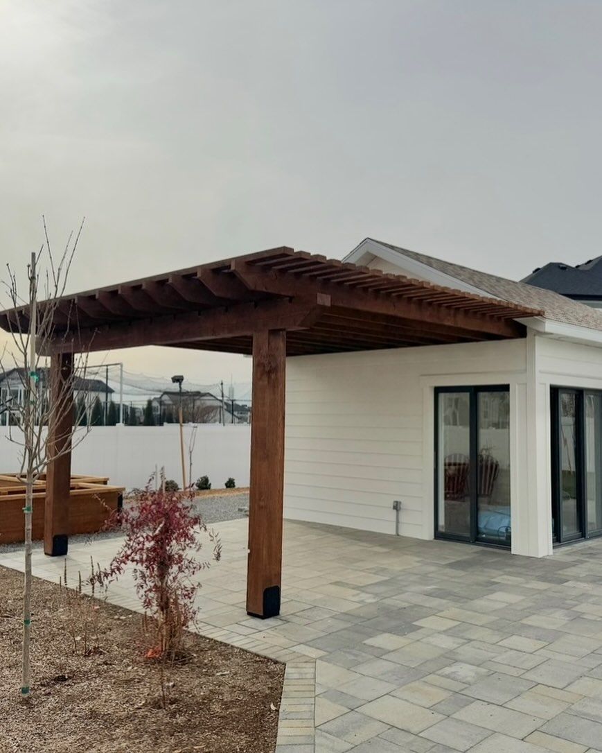 Modern patio with wooden pergola beside a white house and sliding glass doors, on a paved courtyard.