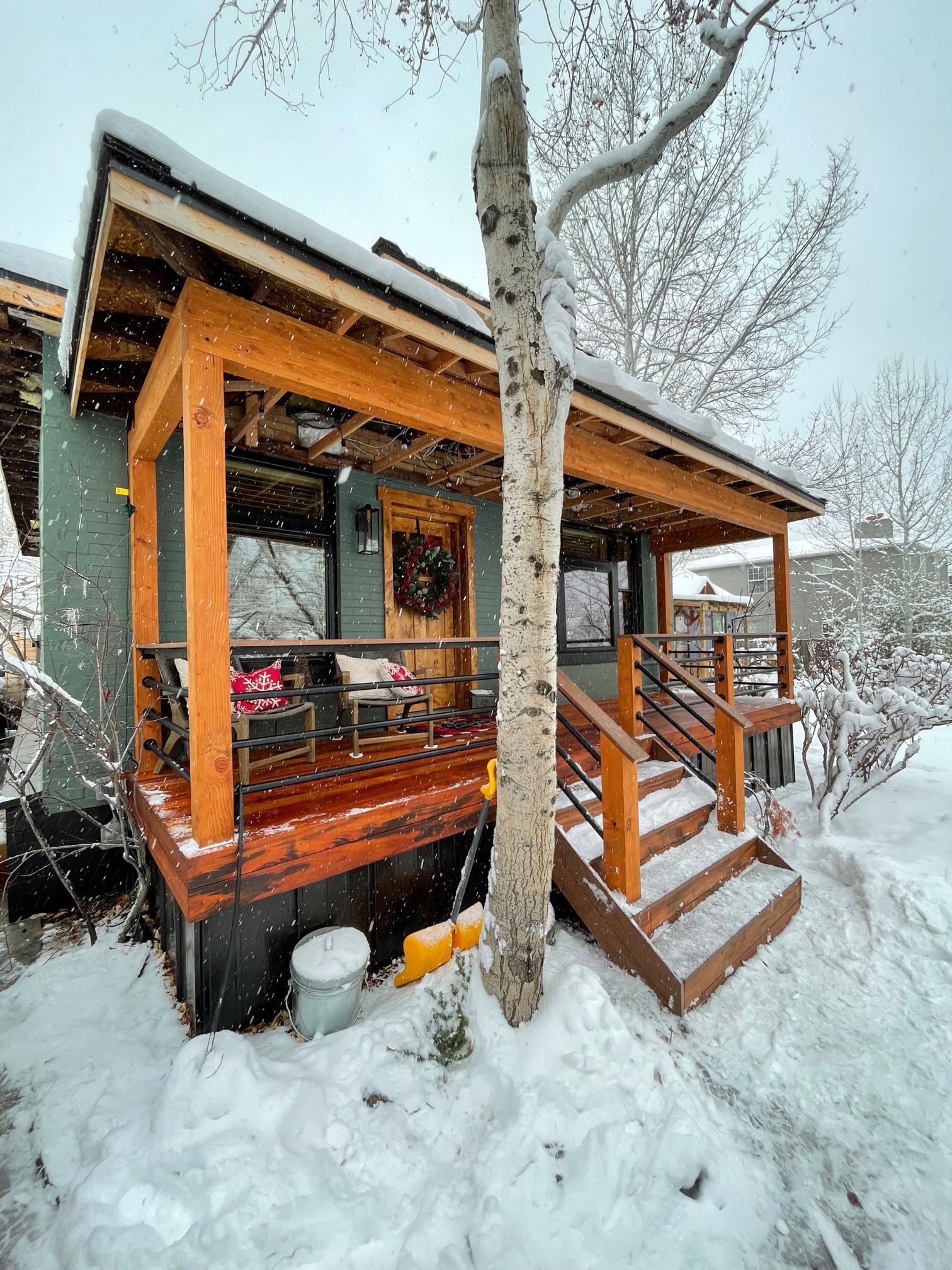 Snow-covered cabin porch with orange-trimmed deck and stairs beside bare trees