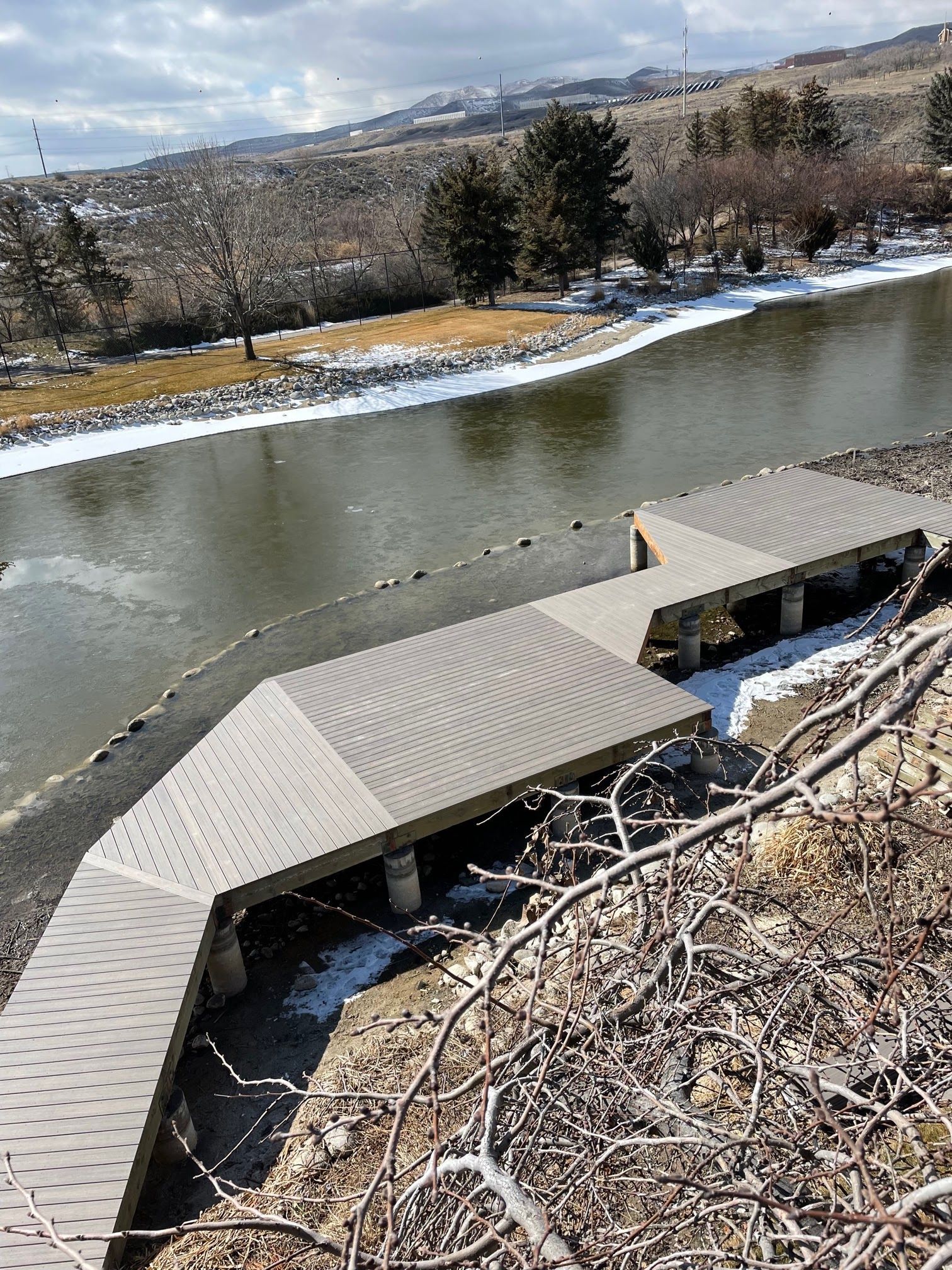 Curved wooden boardwalk along a riverbank with snow and bare trees in a winter landscape