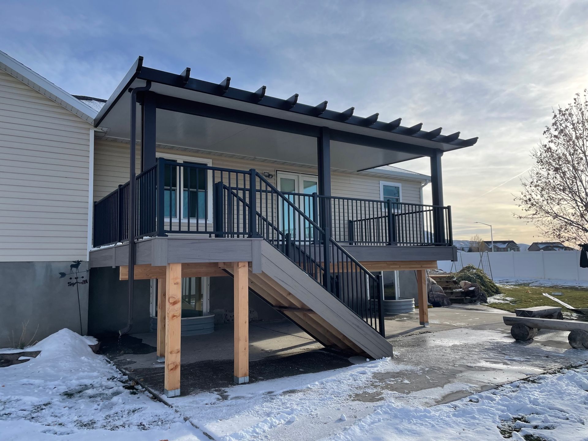 Covered backyard deck with stairs and black railings attached to a house, with snow on the ground