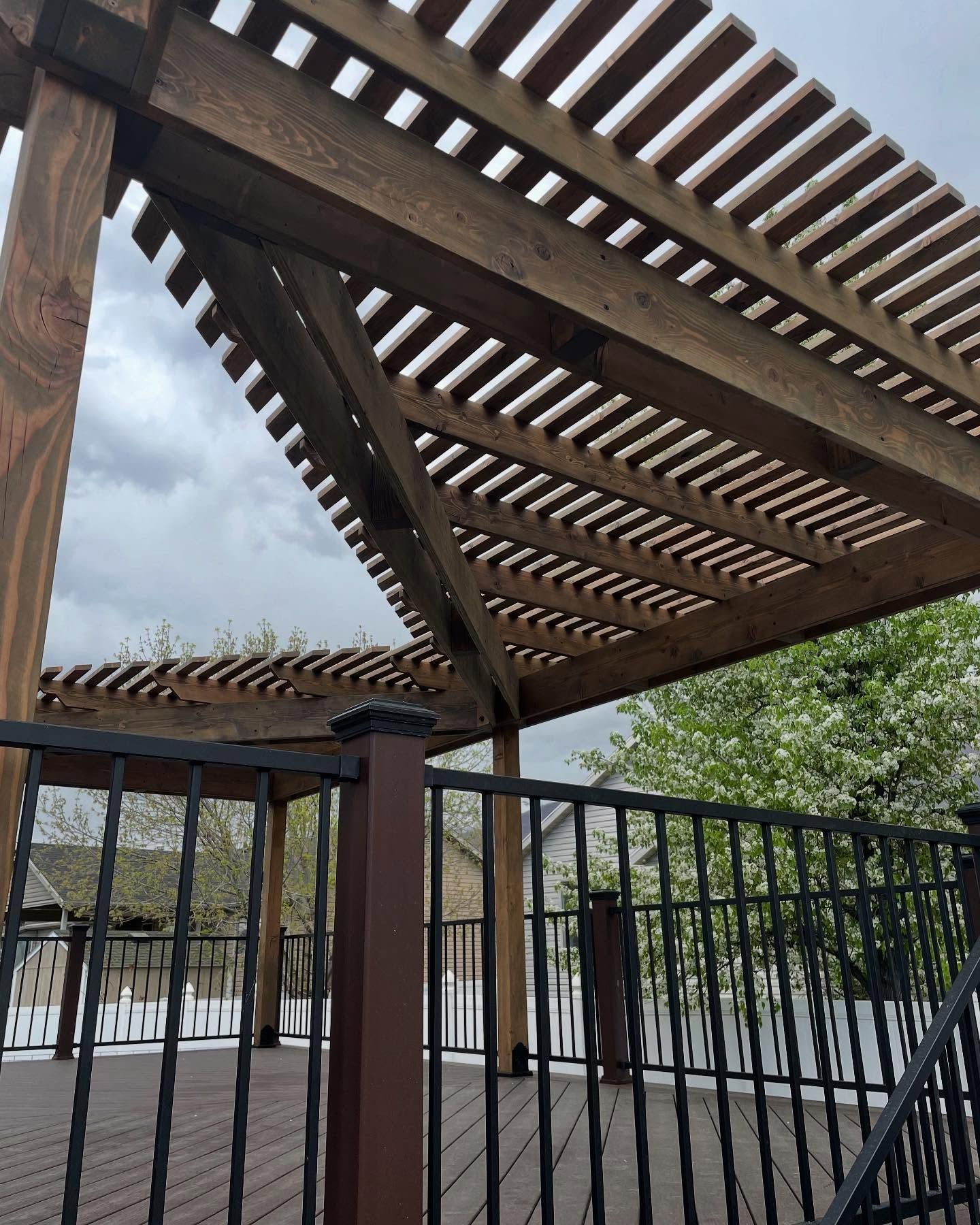 Wooden pergola with slatted roof above a fenced outdoor patio under cloudy sky.