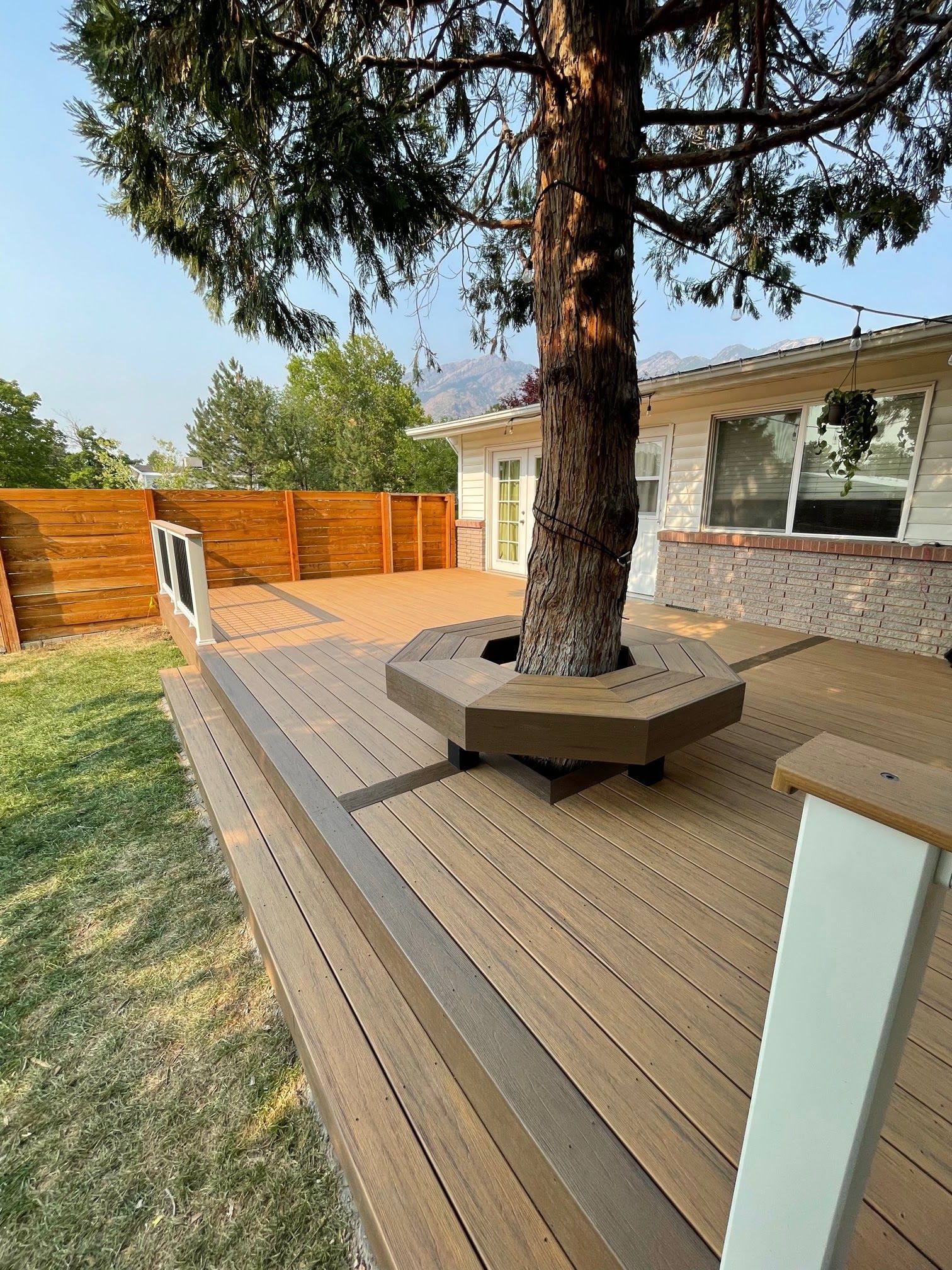 Backyard deck with a tree planter, wooden fencing, and a house exterior with large windows
