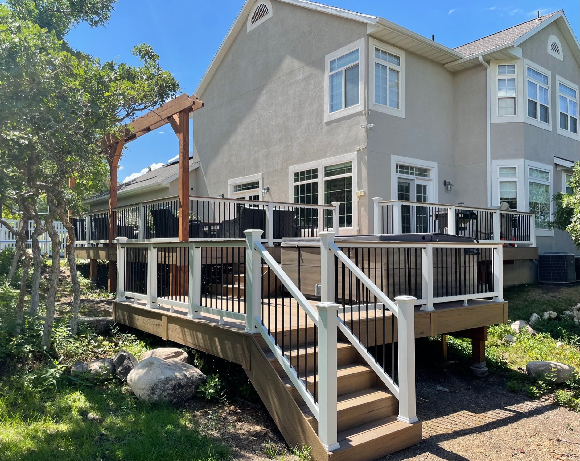 Two-story beige house with a raised wooden deck and stairs in a sunny yard