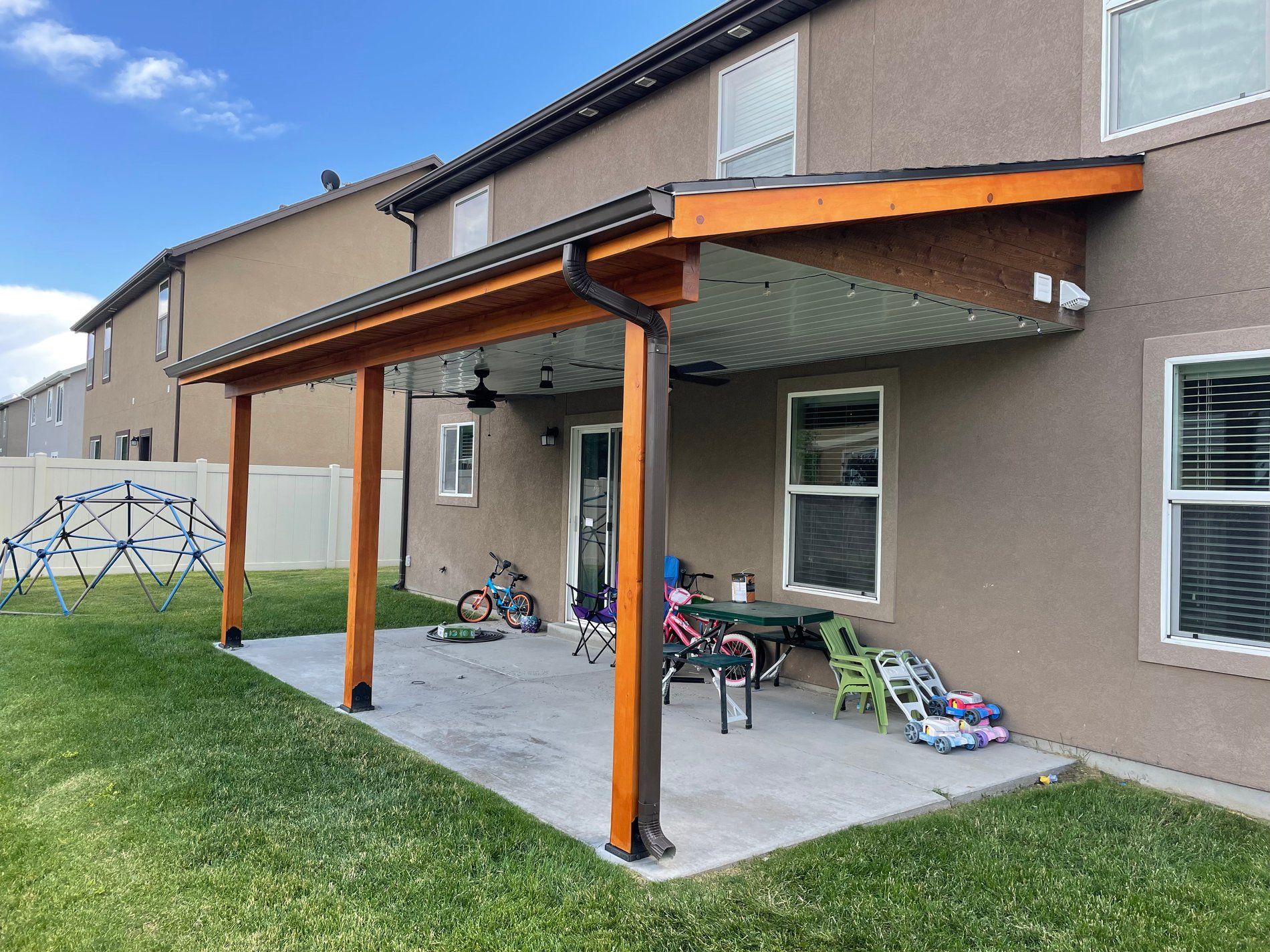 Back patio with a wooden pergola, patio furniture, bikes, and a grassy yard beside a house.
