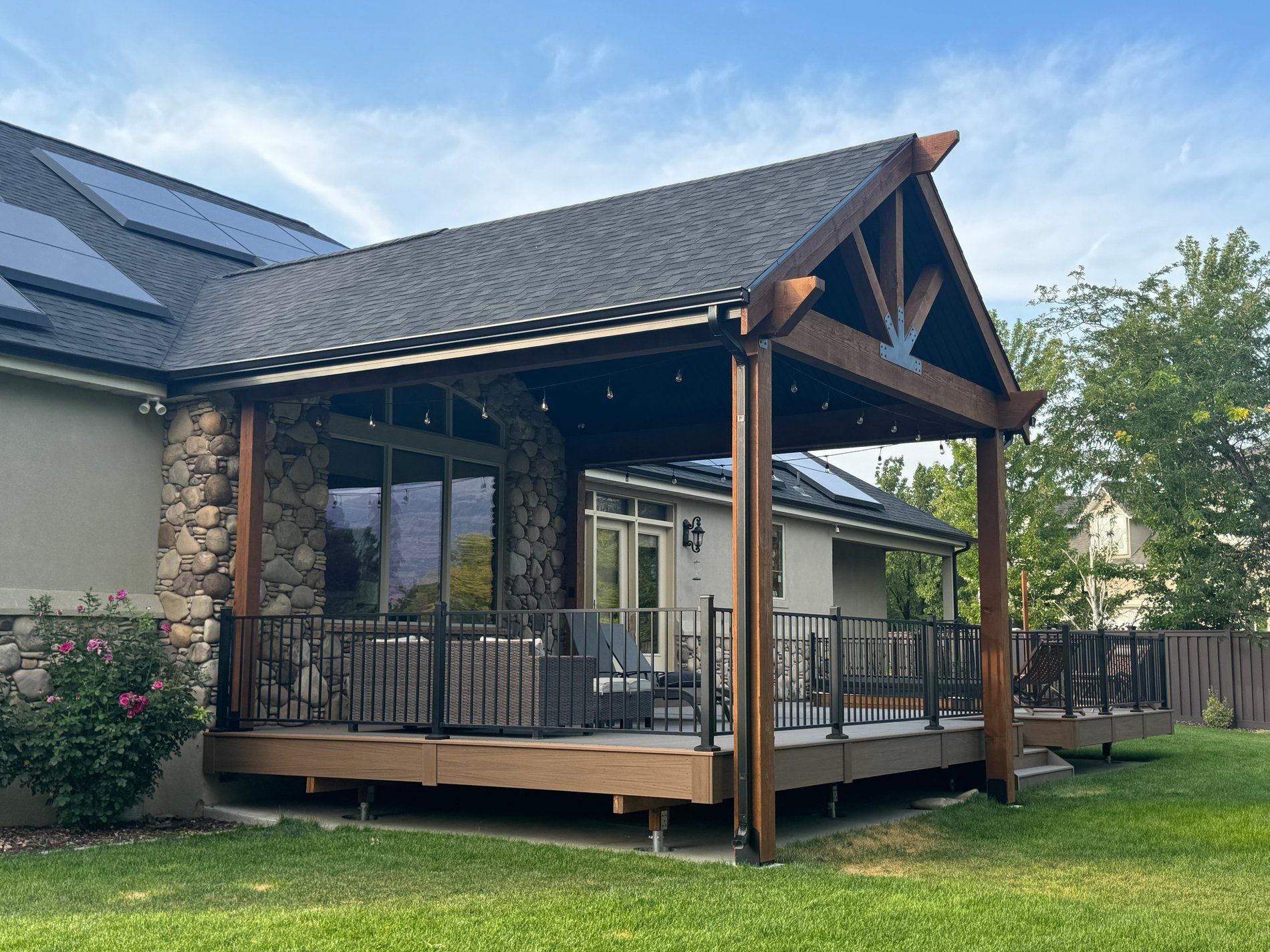 Covered wooden patio deck with railing beside a stone house, under a pitched roof in a grassy yard