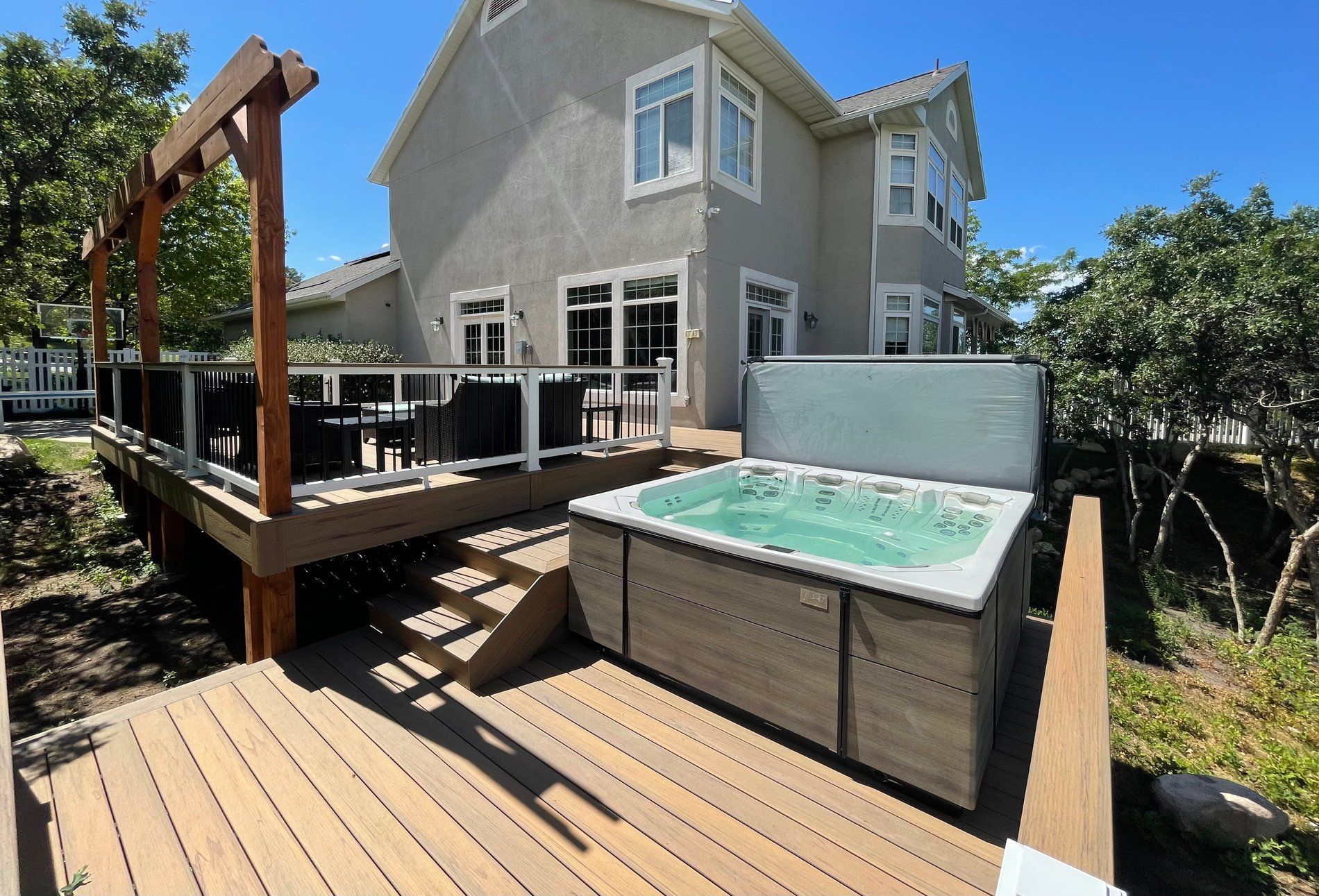 Backyard deck with hot tub beside a gray house and wooden stairs under a blue sky