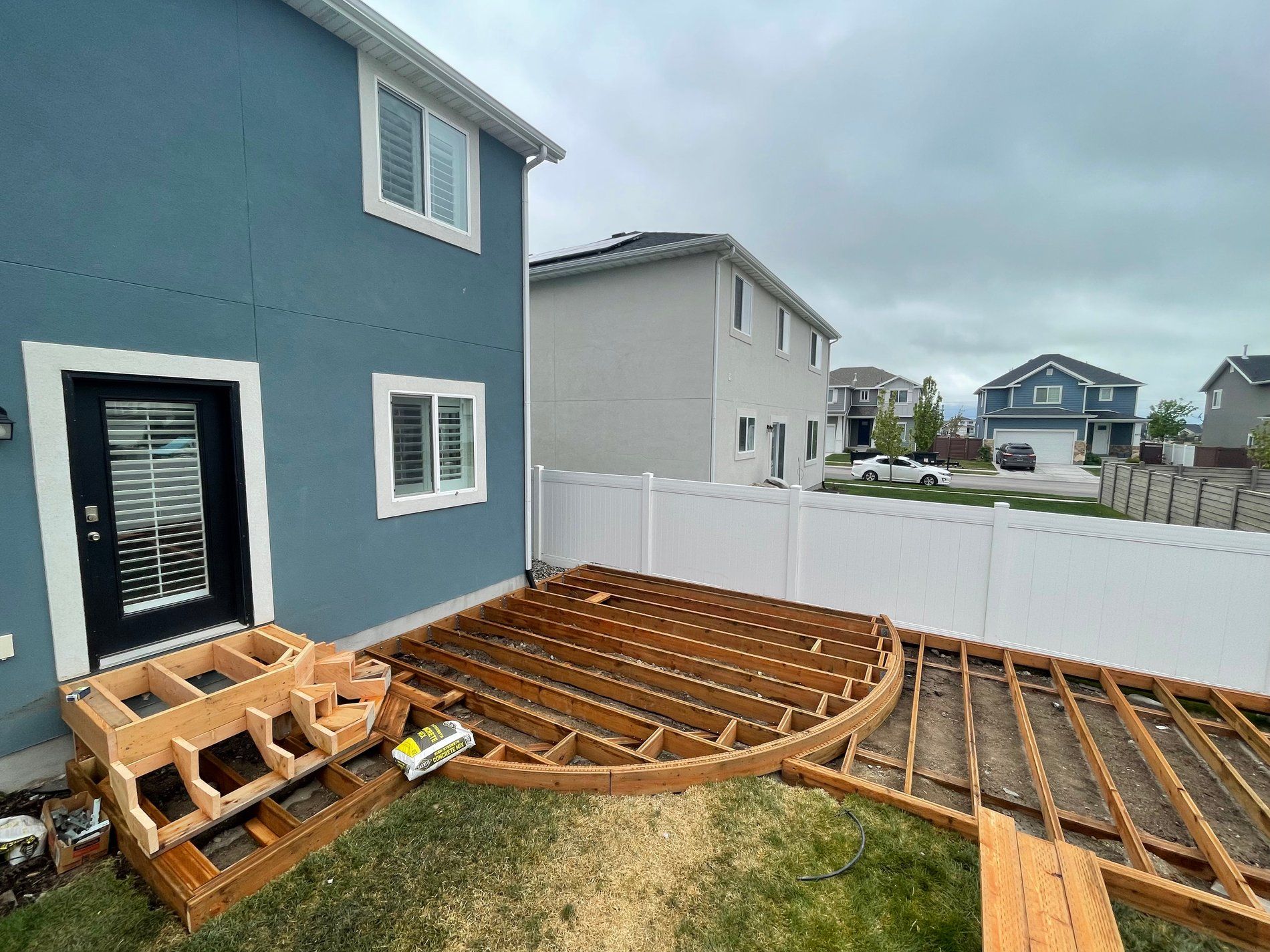 Backyard deck under construction beside a blue house, with wooden framing and white fence.
