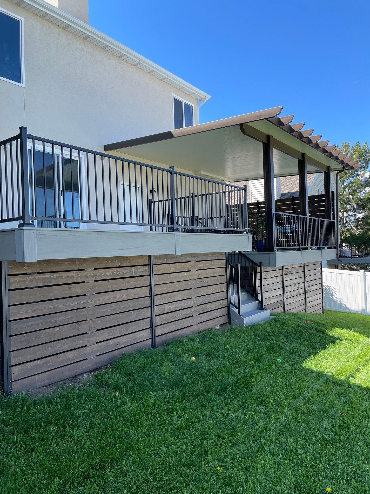 Raised wooden deck with black railing and covered seating area beside a house, overlooking a green lawn
