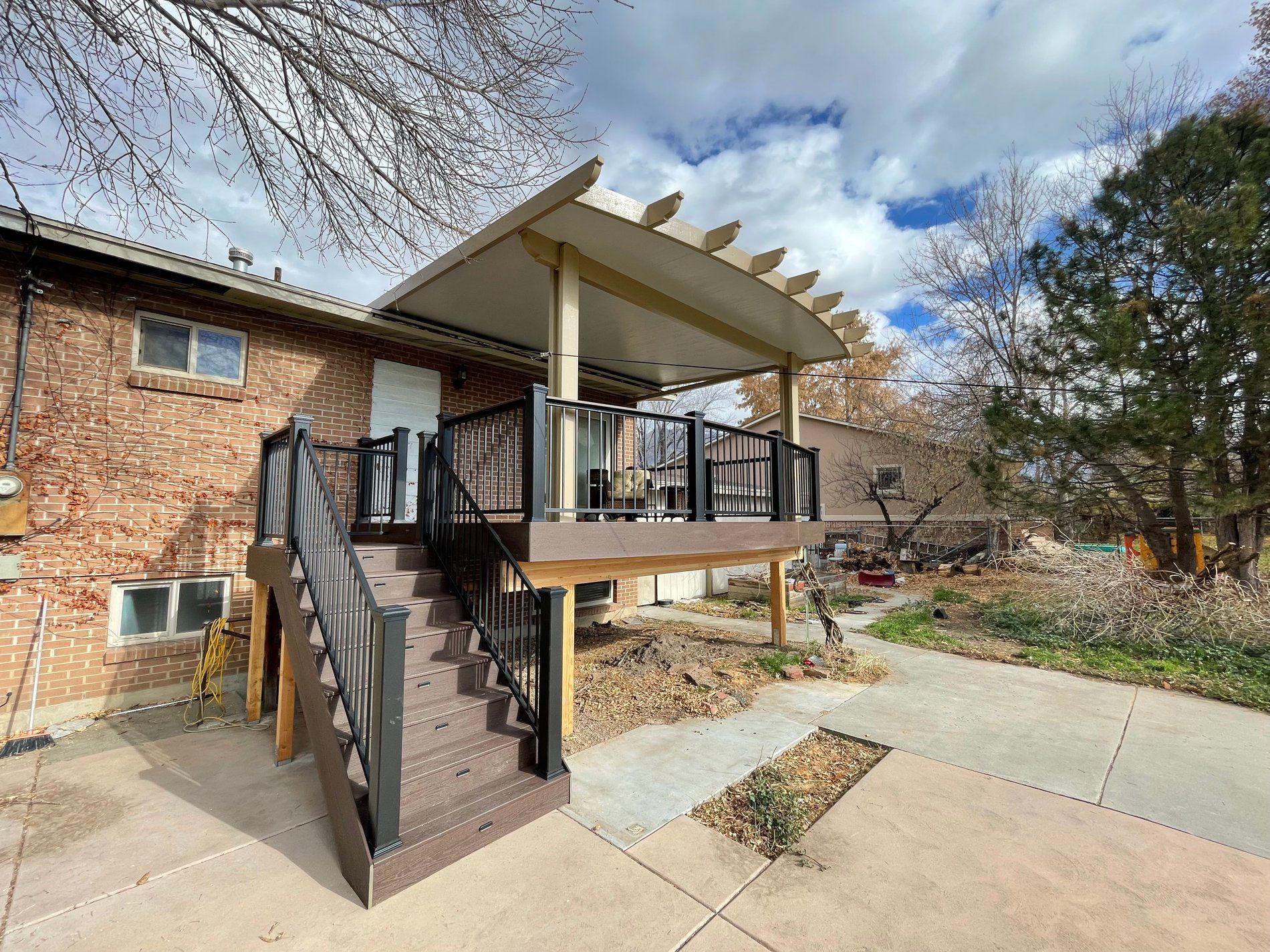 Brick house with a small elevated deck, black stairs, and a concrete driveway under a cloudy sky
