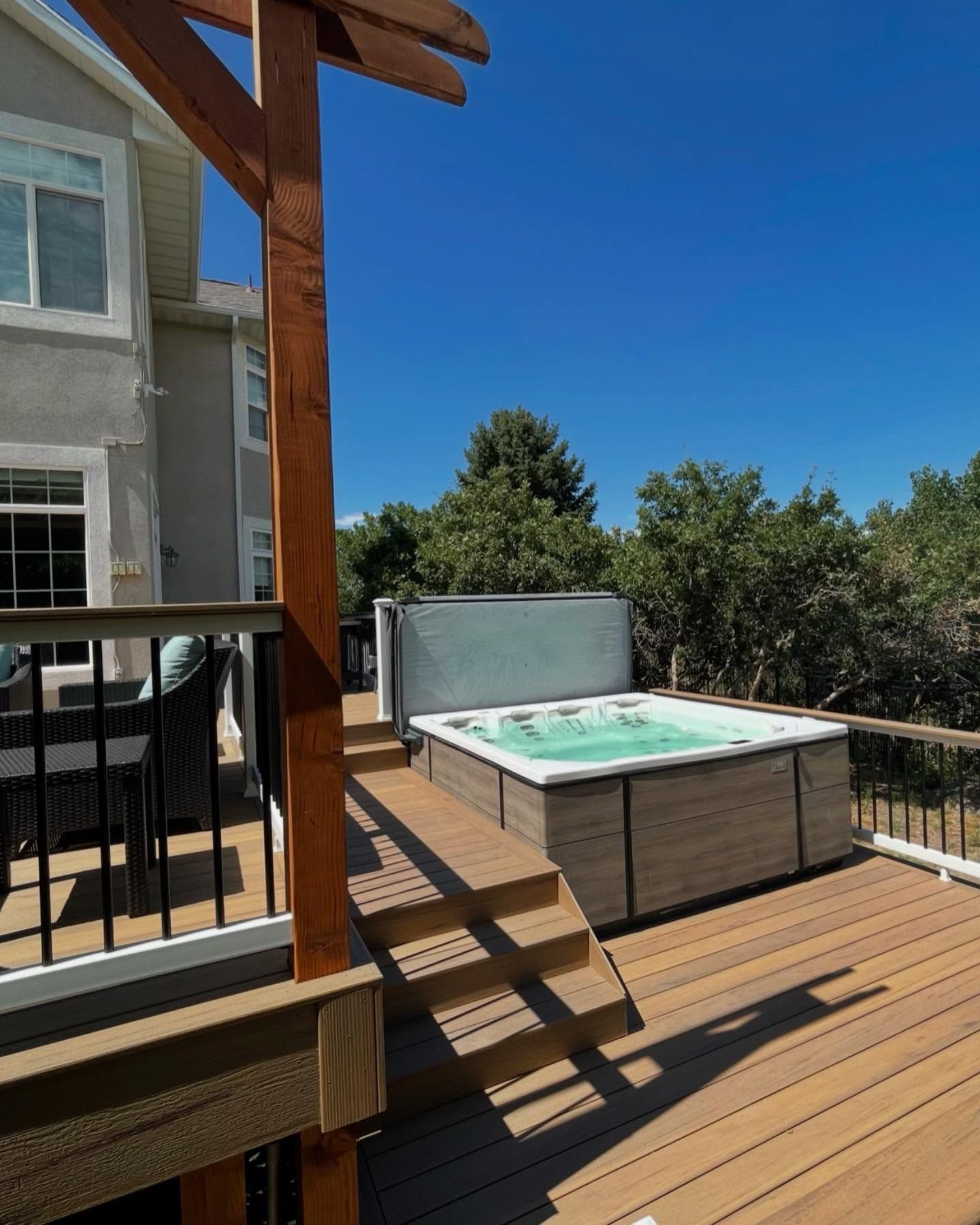 Wooden deck with a hot tub beside a house, framed by black railing and trees under a clear blue sky