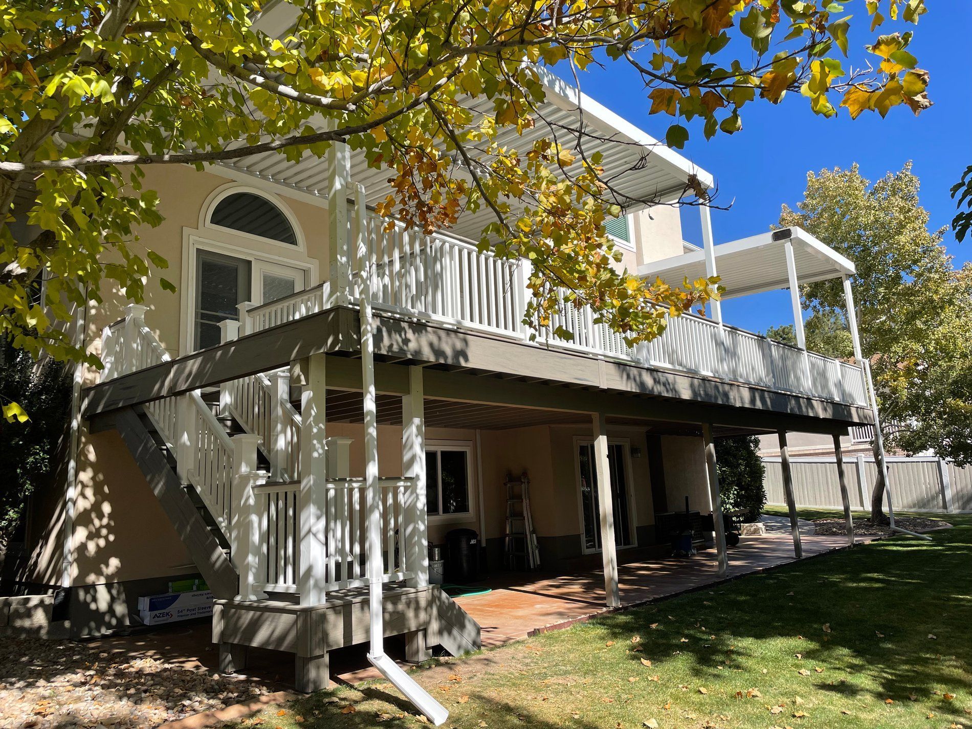 Two-story house with a covered deck, stairs, and yard under a blue sky, framed by autumn trees