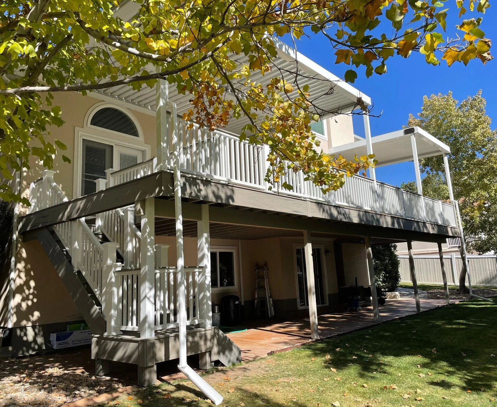 Two-story cream house with a white balcony and shaded ground-level patio under leafy trees