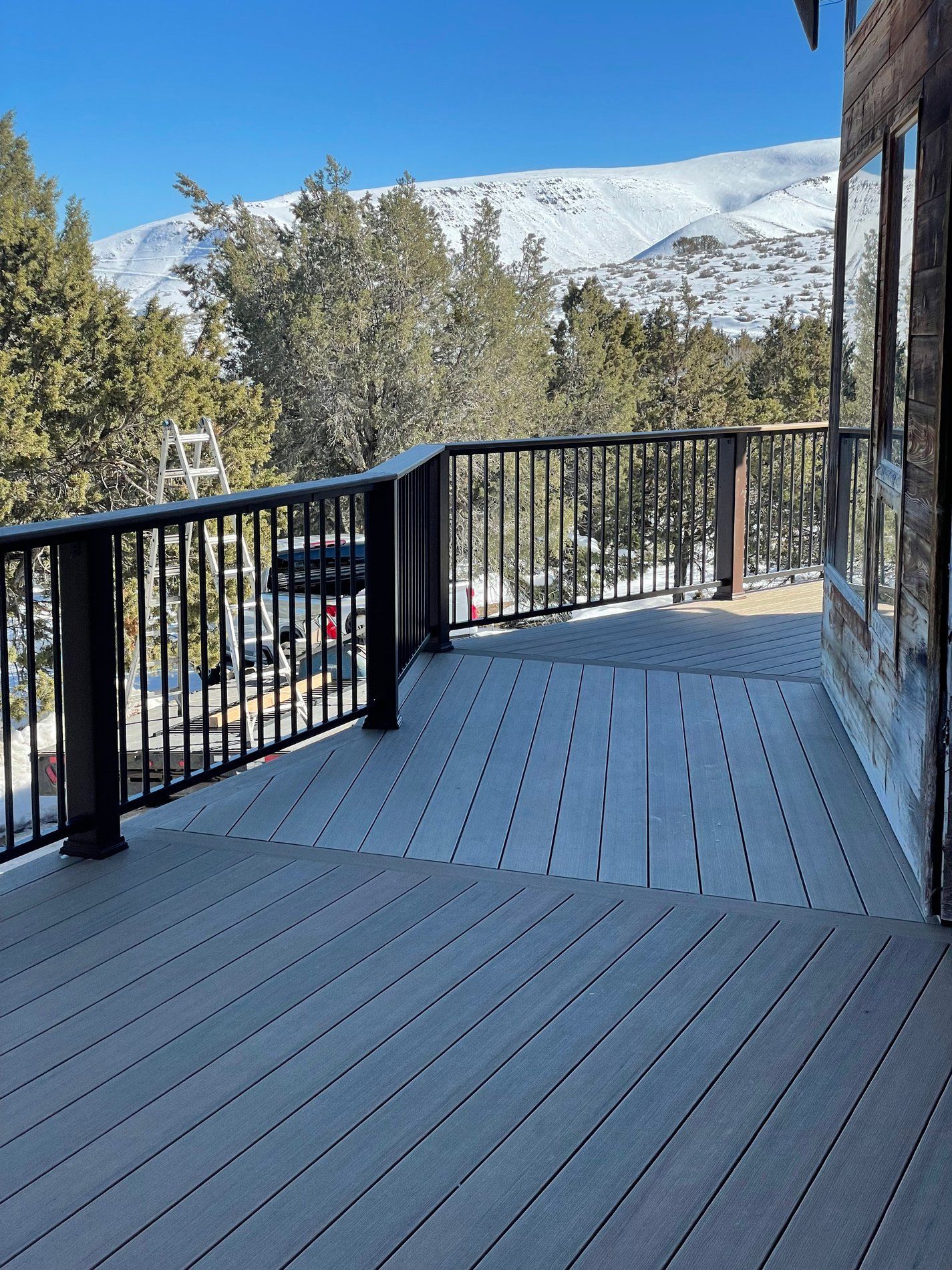 Snowy mountain view from a wooden deck with black railing and trees under a blue sky