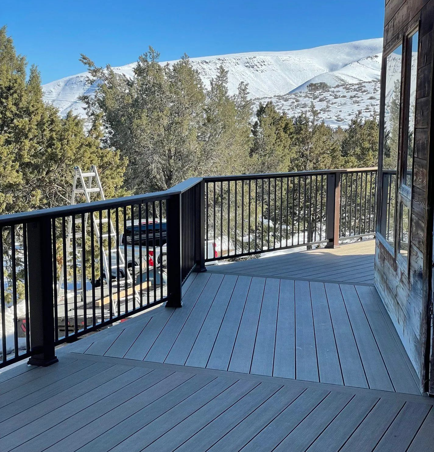 Snowy mountain view from a deck with black railing and trees under a clear blue sky