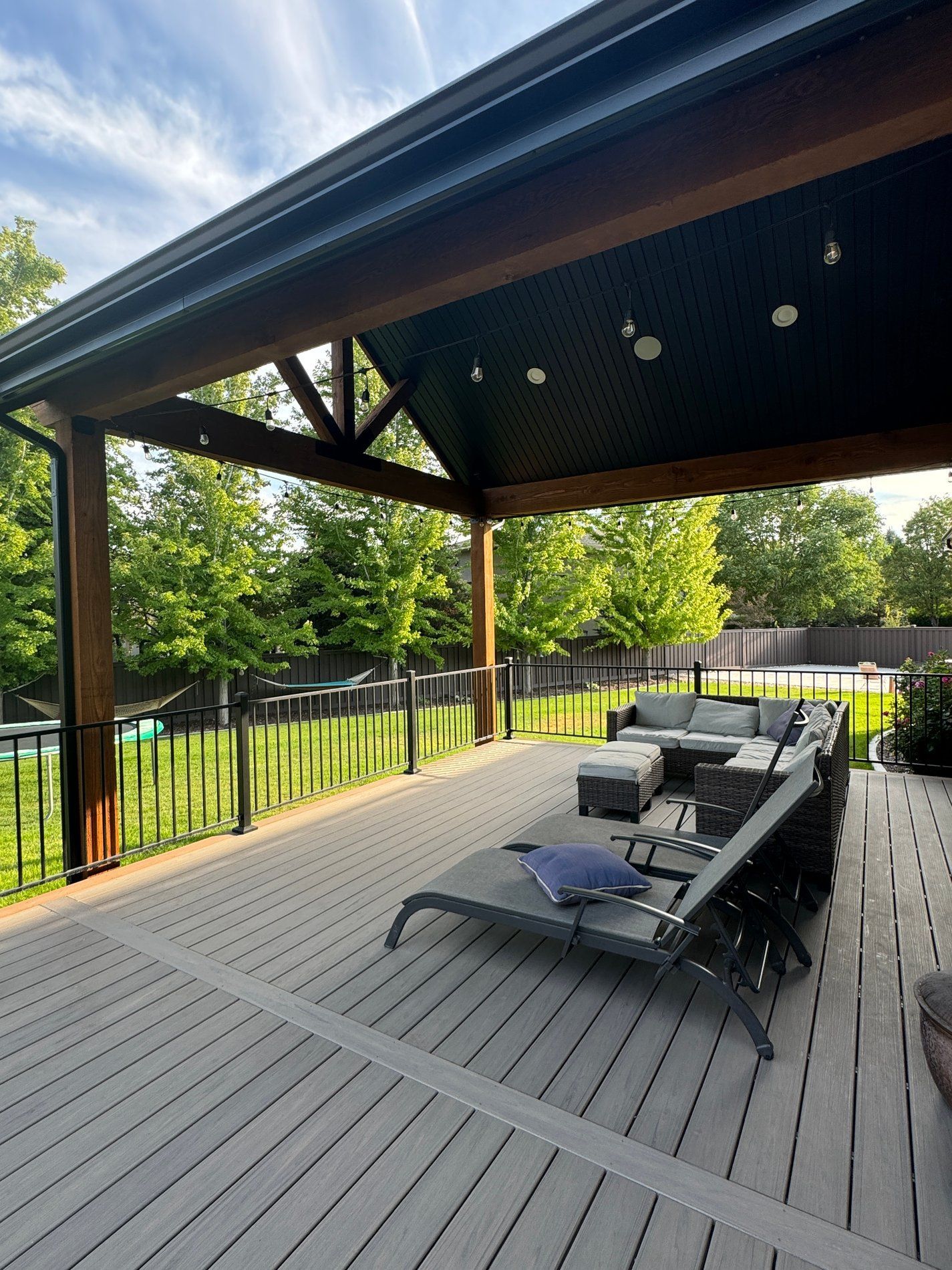Covered wooden deck with lounge chairs, black railing, and green trees beyond a yard fence