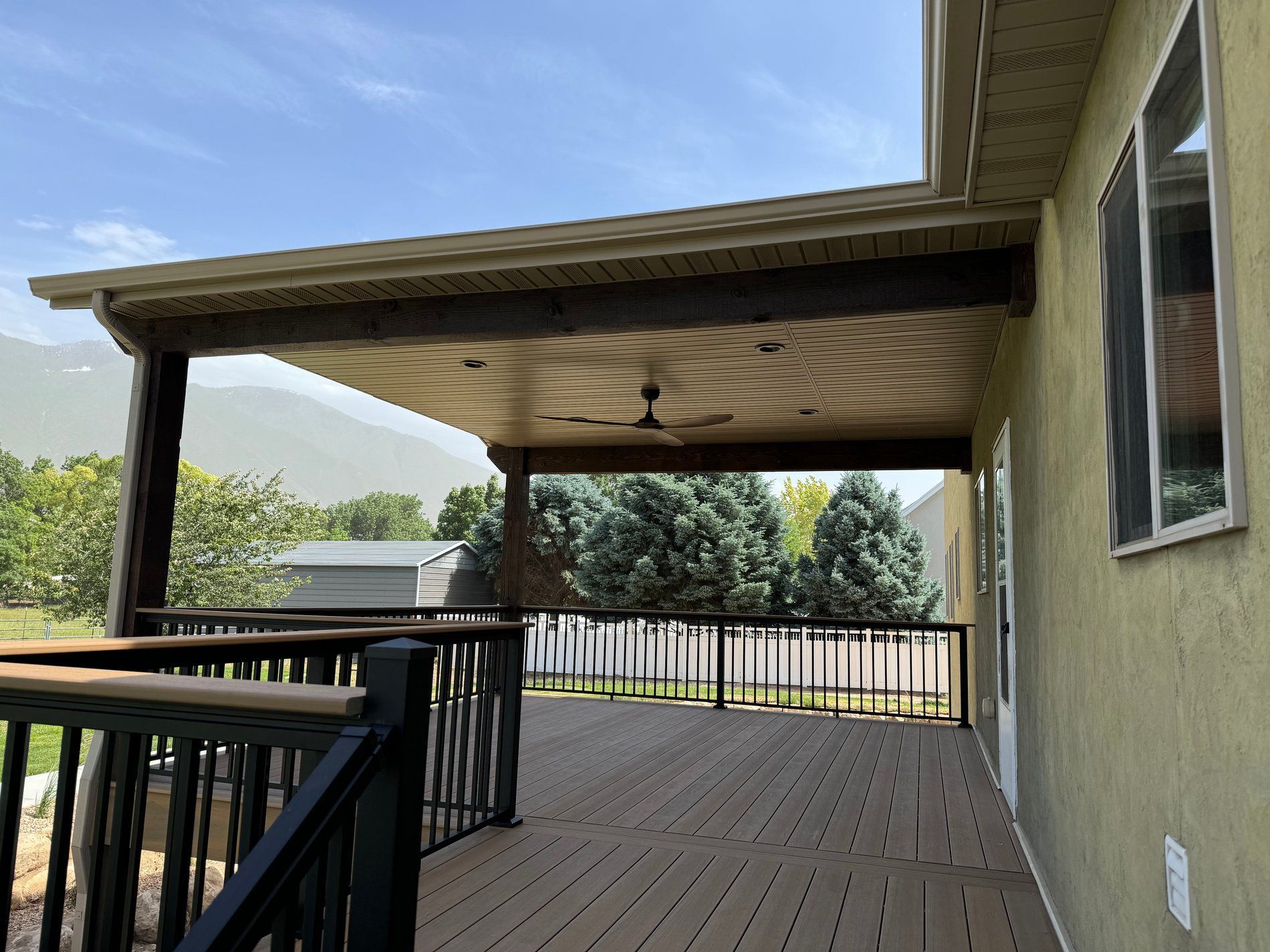 Covered wooden deck with railing beside a beige house, overlooking trees and a distant field under a blue sky