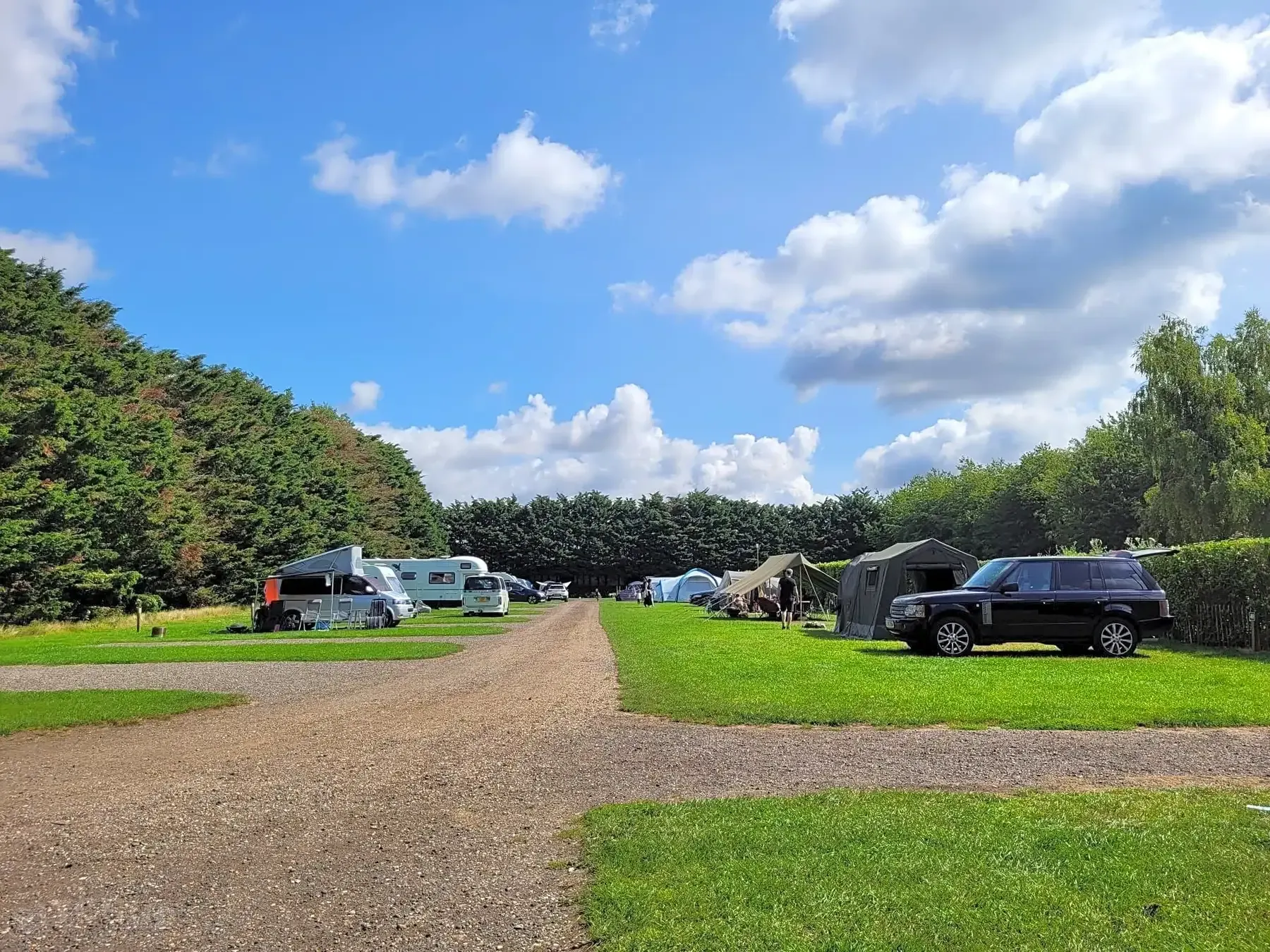 Campsite with RVs, tents, and vehicles on green grass, under a blue sky with white clouds.
