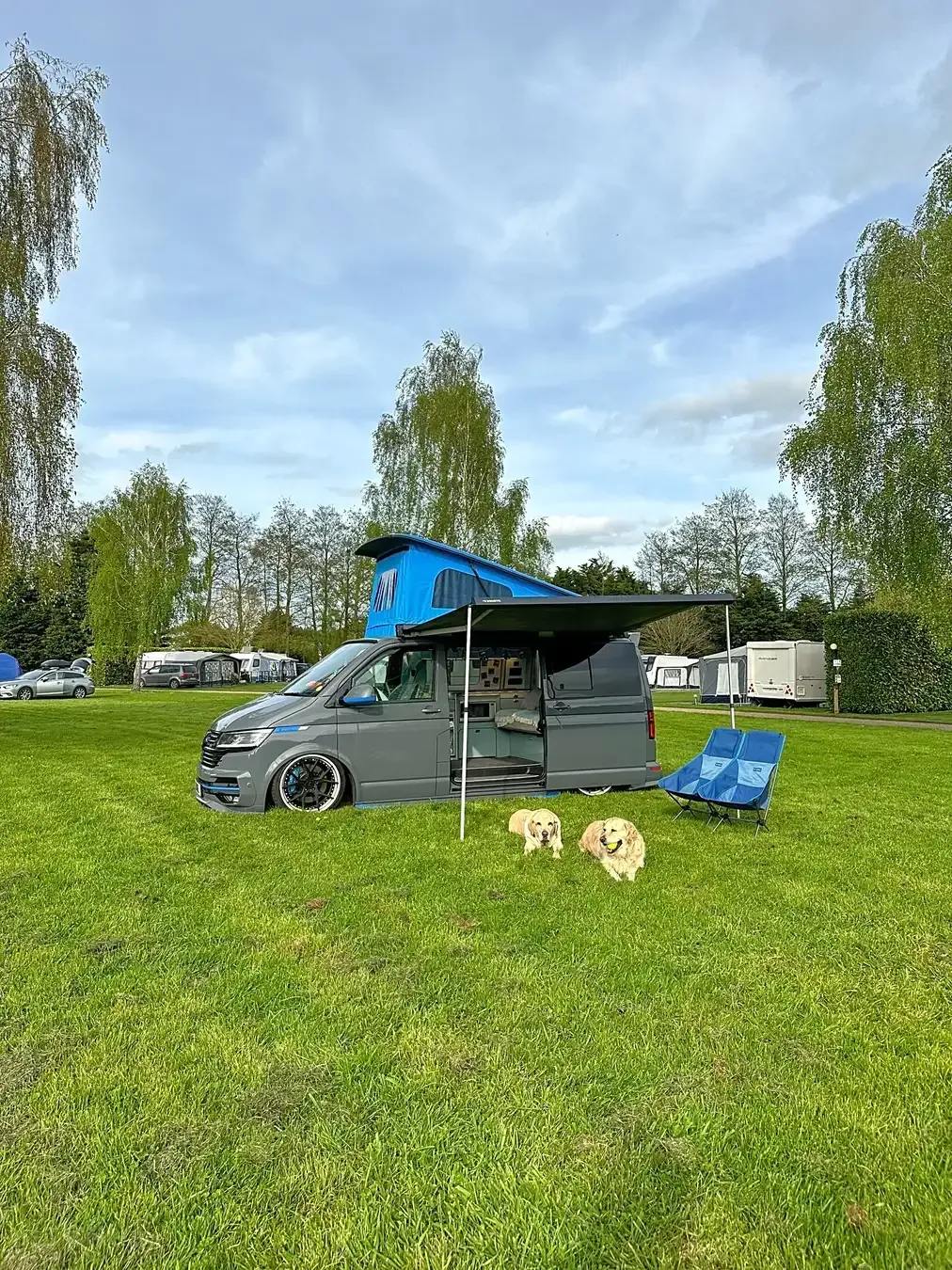 Gray camper van with pop-up roof and awning on a grassy campsite with two dogs and blue folding chair.