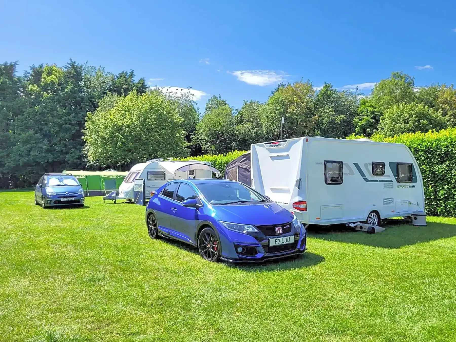 Blue car and two caravans parked on a green lawn with trees in the background under a blue sky.