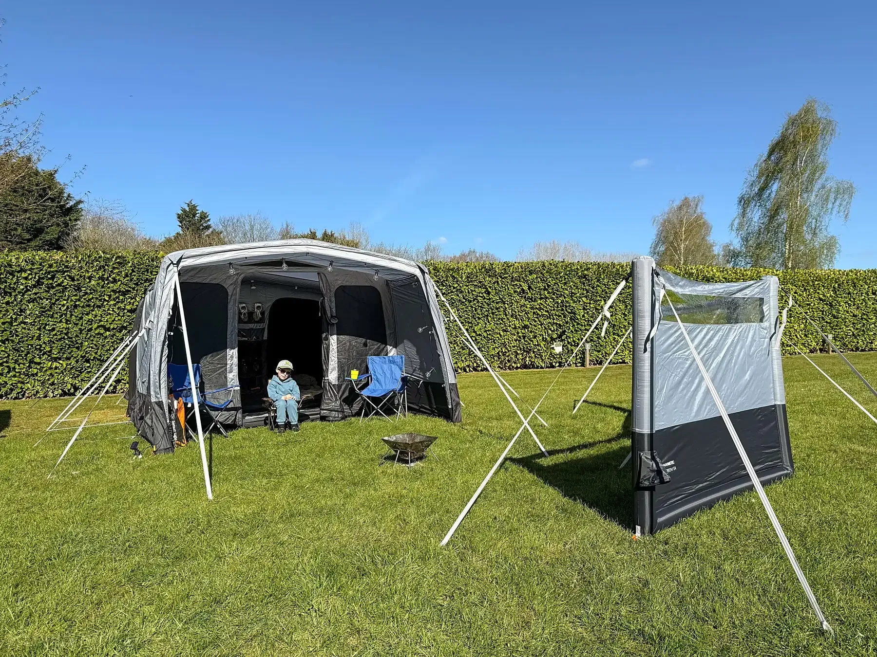 Van with awning set up for camping on green grass. Two figures sit inside. A screen stands beside it.