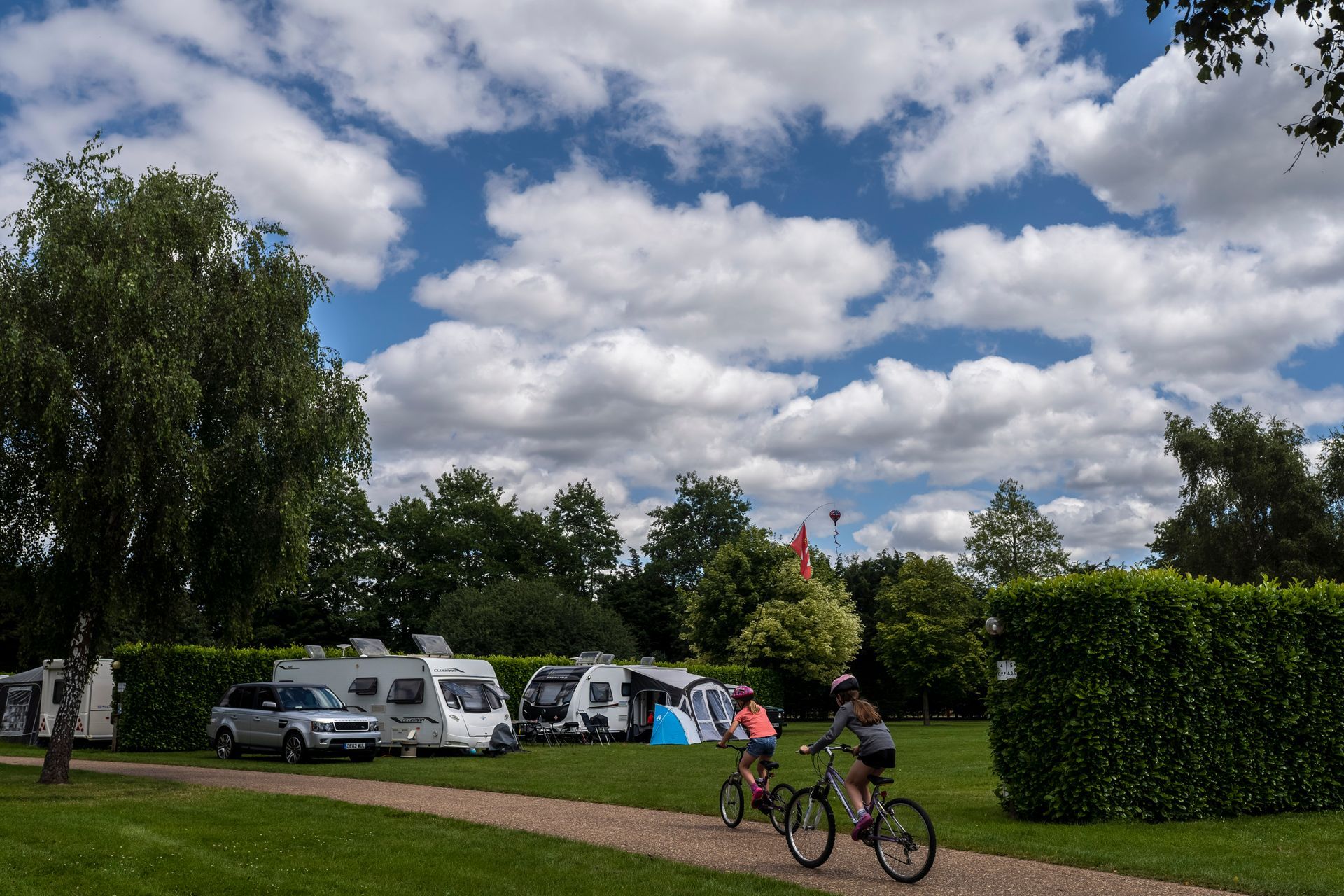 Campground with trailers, cyclists, green lawn, trees, and cloudy blue sky.